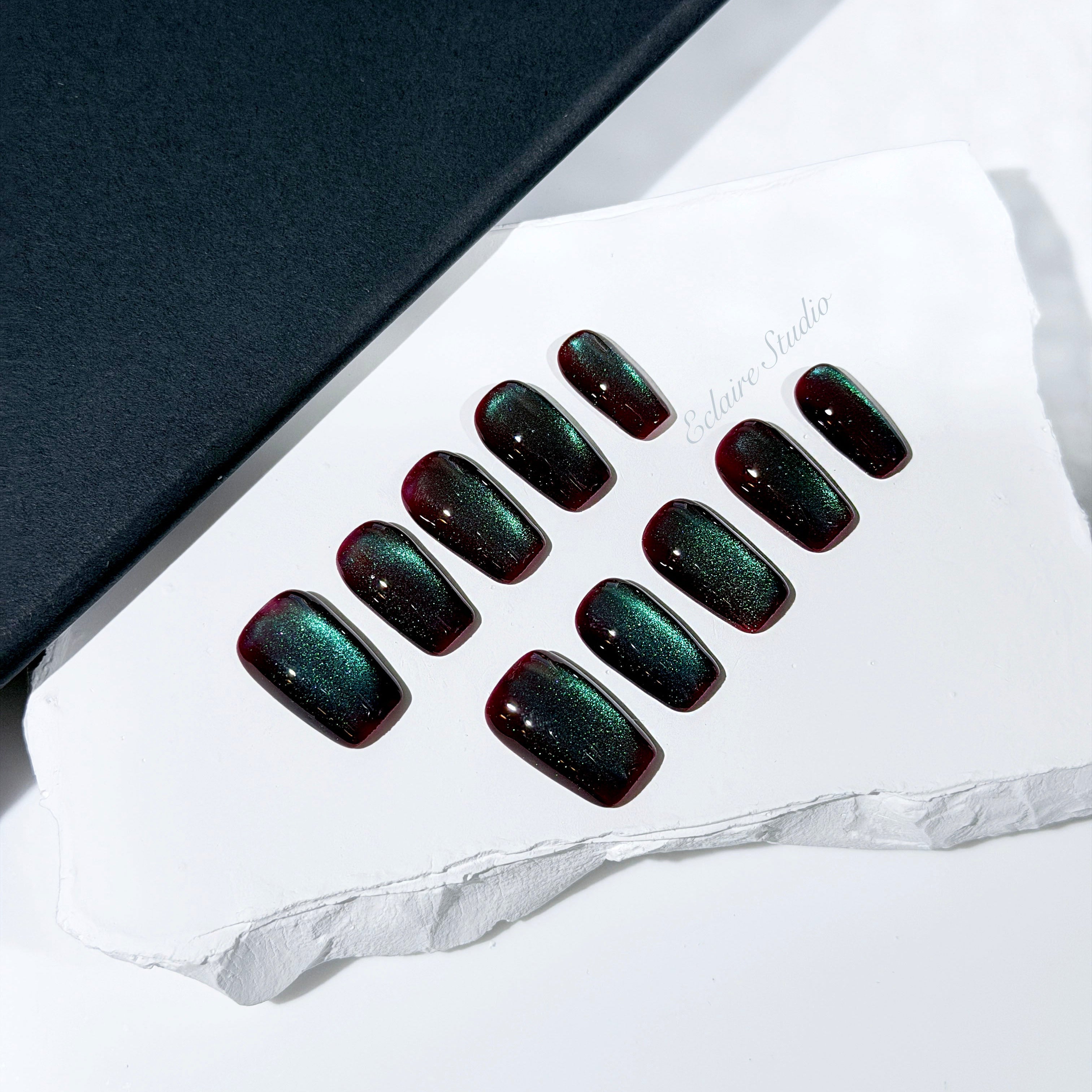 Medium coffin press-on nails with deep cherry-black base and aurora green cat-eye shimmer, arranged on a white stone surface.