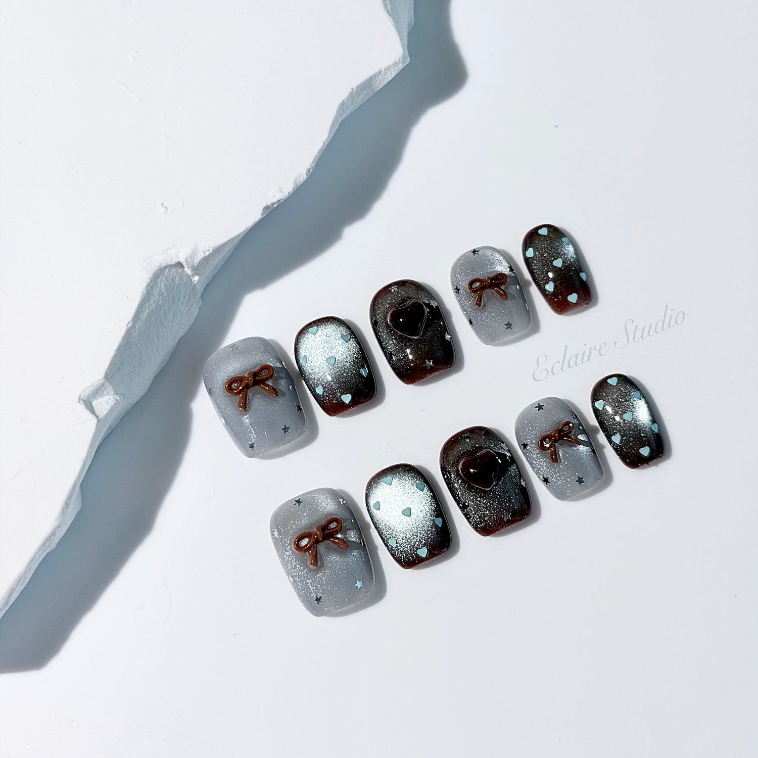 Short coffin press-on nails in mint and chocolate brown gradient with 3D heart and bow accents, displayed in a clear Éclaire Studio case under soft lighting.