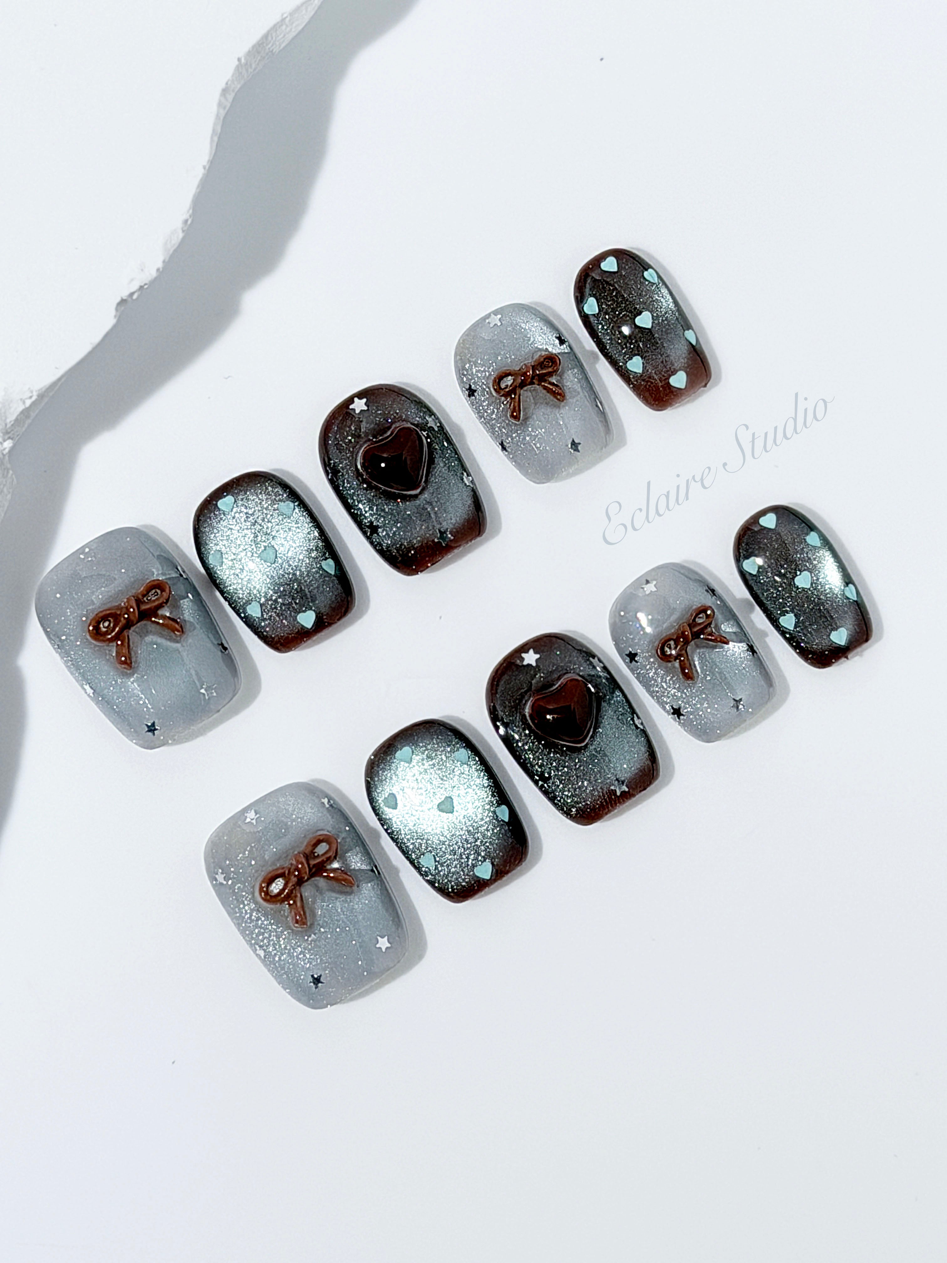 Short coffin press-on nails in mint and chocolate brown gradient with 3D heart and bow accents, displayed in a clear Éclaire Studio case under soft lighting.