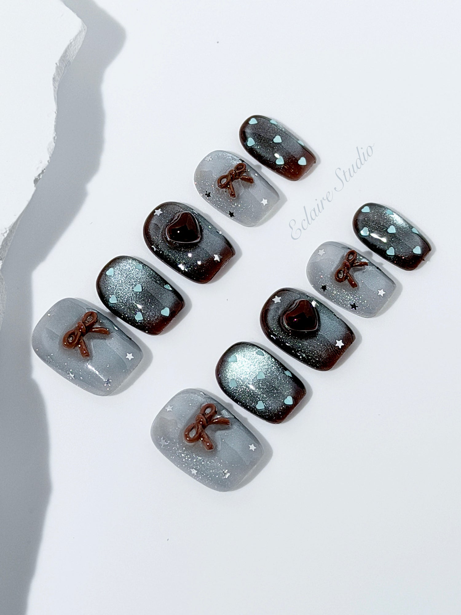 Short coffin press-on nails in mint and chocolate brown gradient with 3D heart and bow accents, displayed in a clear Éclaire Studio case under soft lighting.