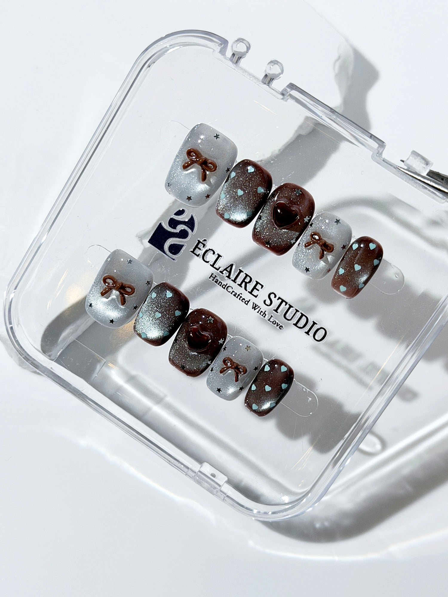 Short coffin press-on nails in mint and chocolate brown gradient with 3D heart and bow accents, displayed in a clear Éclaire Studio case under soft lighting.