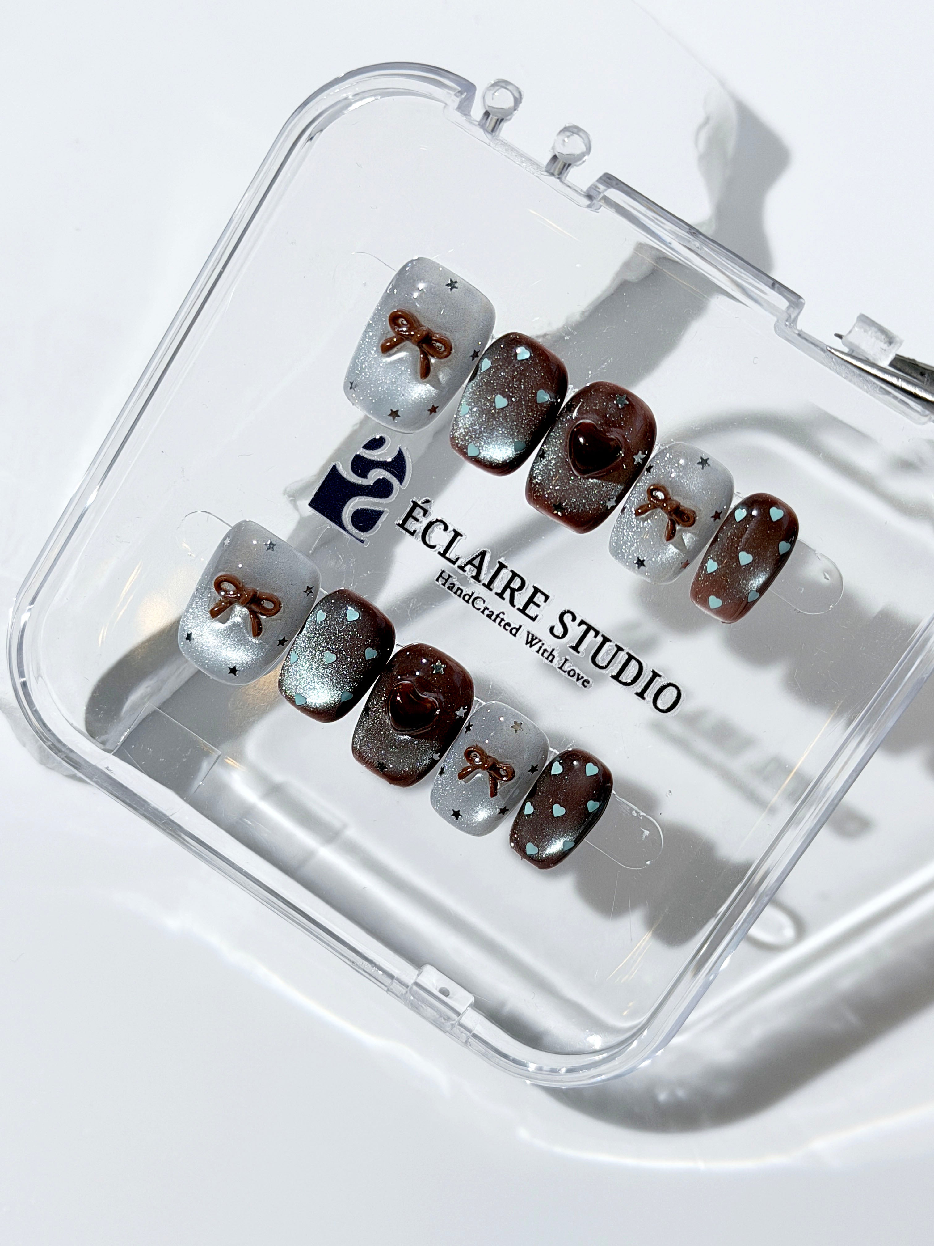 Short coffin press-on nails in mint and chocolate brown gradient with 3D heart and bow accents, displayed in a clear Éclaire Studio case under soft lighting.