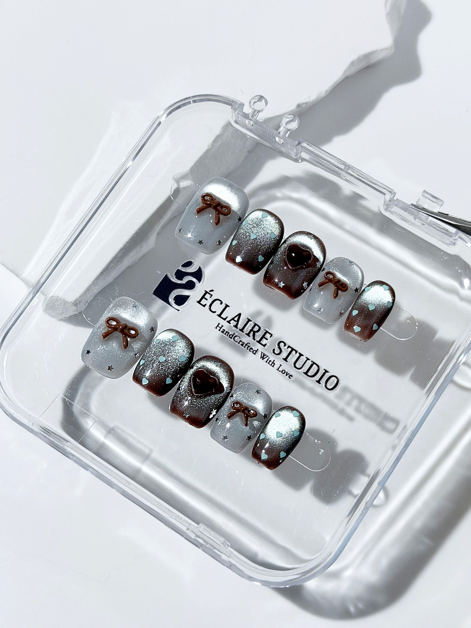 Short coffin press-on nails in mint and chocolate brown gradient with 3D heart and bow accents, displayed in a clear Éclaire Studio case under soft lighting.