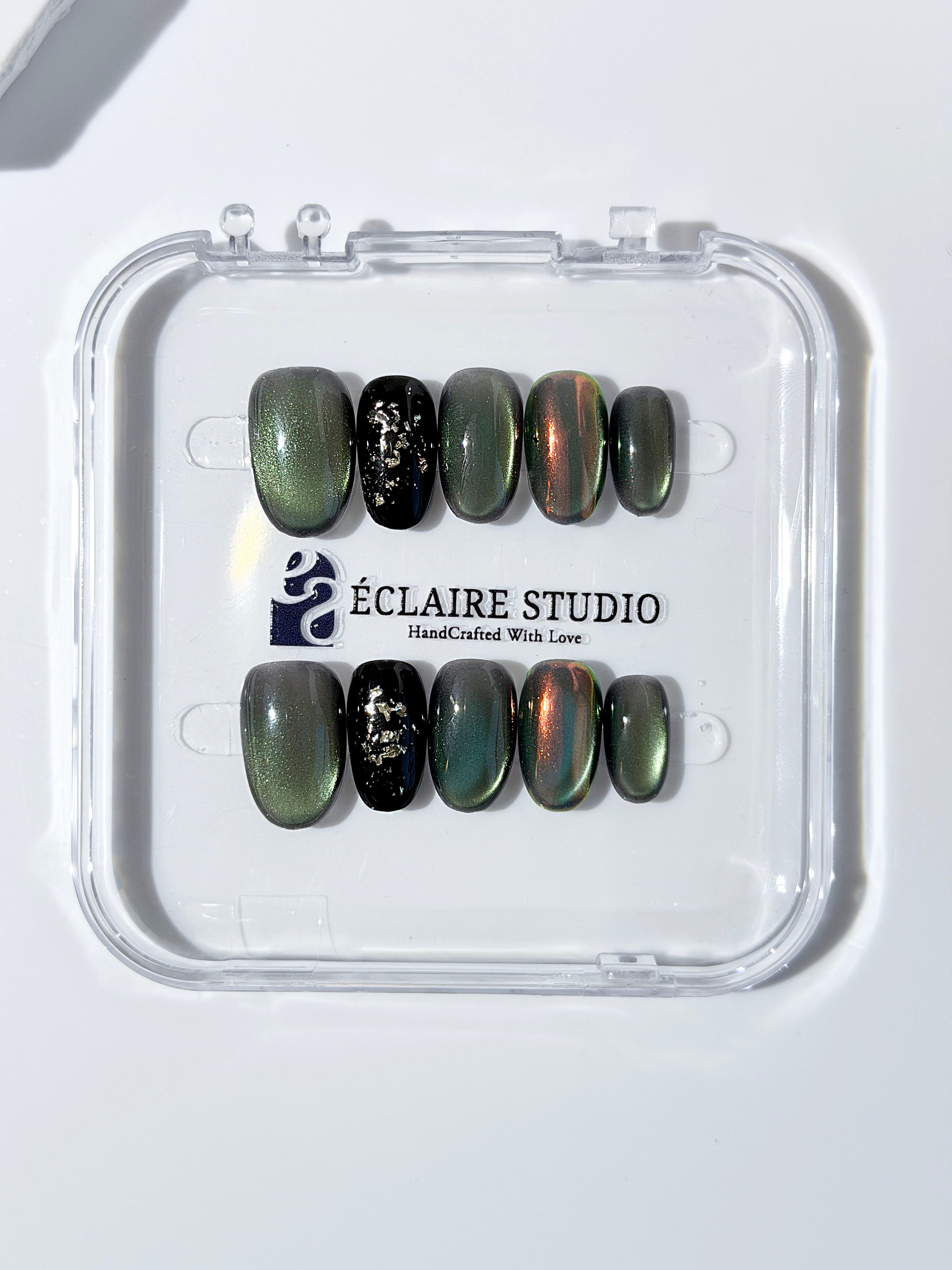 Short round press-on nails in shifting evergreen cat-eye shimmer with black and gold foil accent, displayed in a clear Éclaire Studio case.
