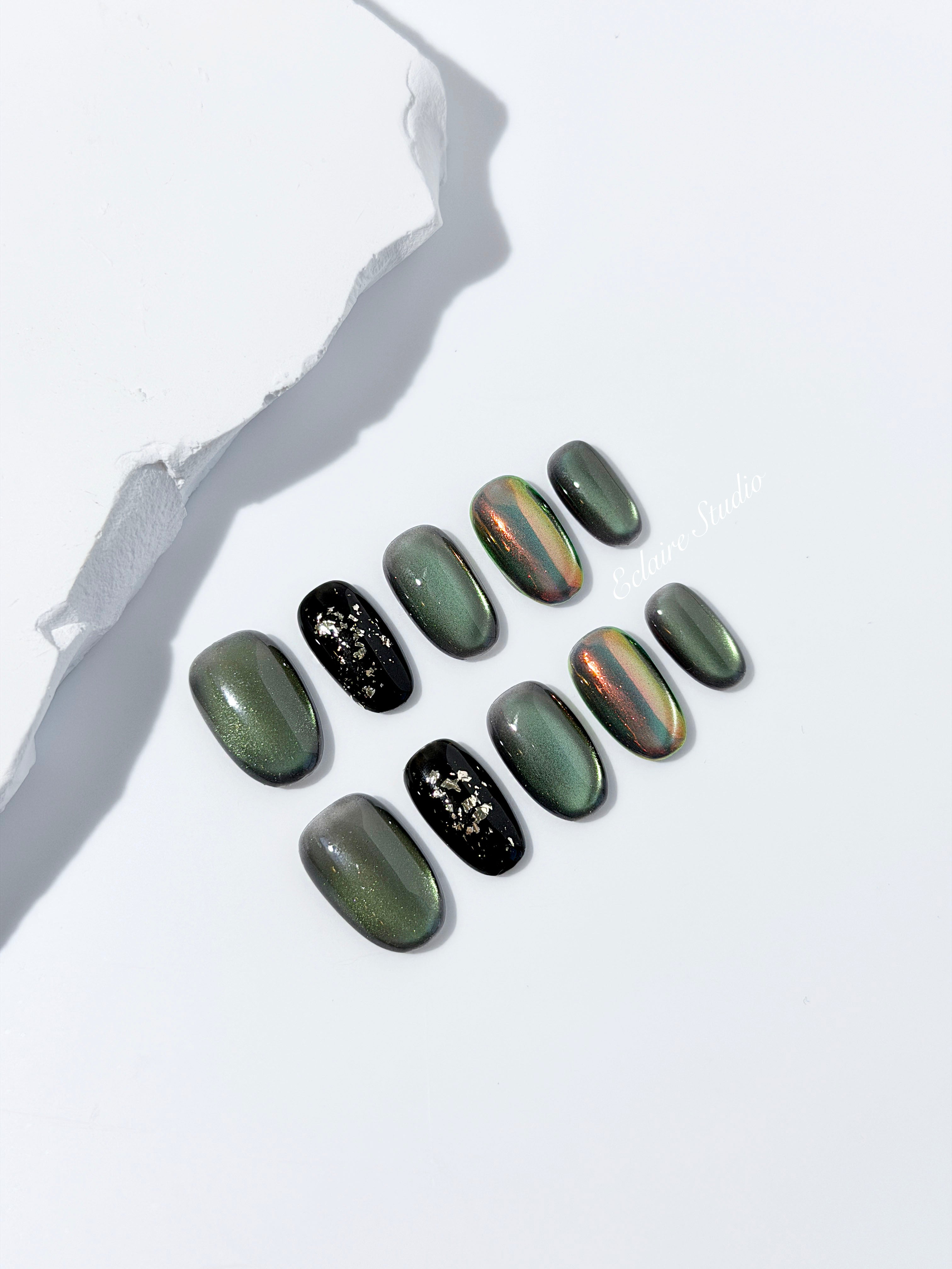Short round press-on nails in shifting evergreen cat-eye shimmer with black and gold foil accent, displayed in a clear Éclaire Studio case.