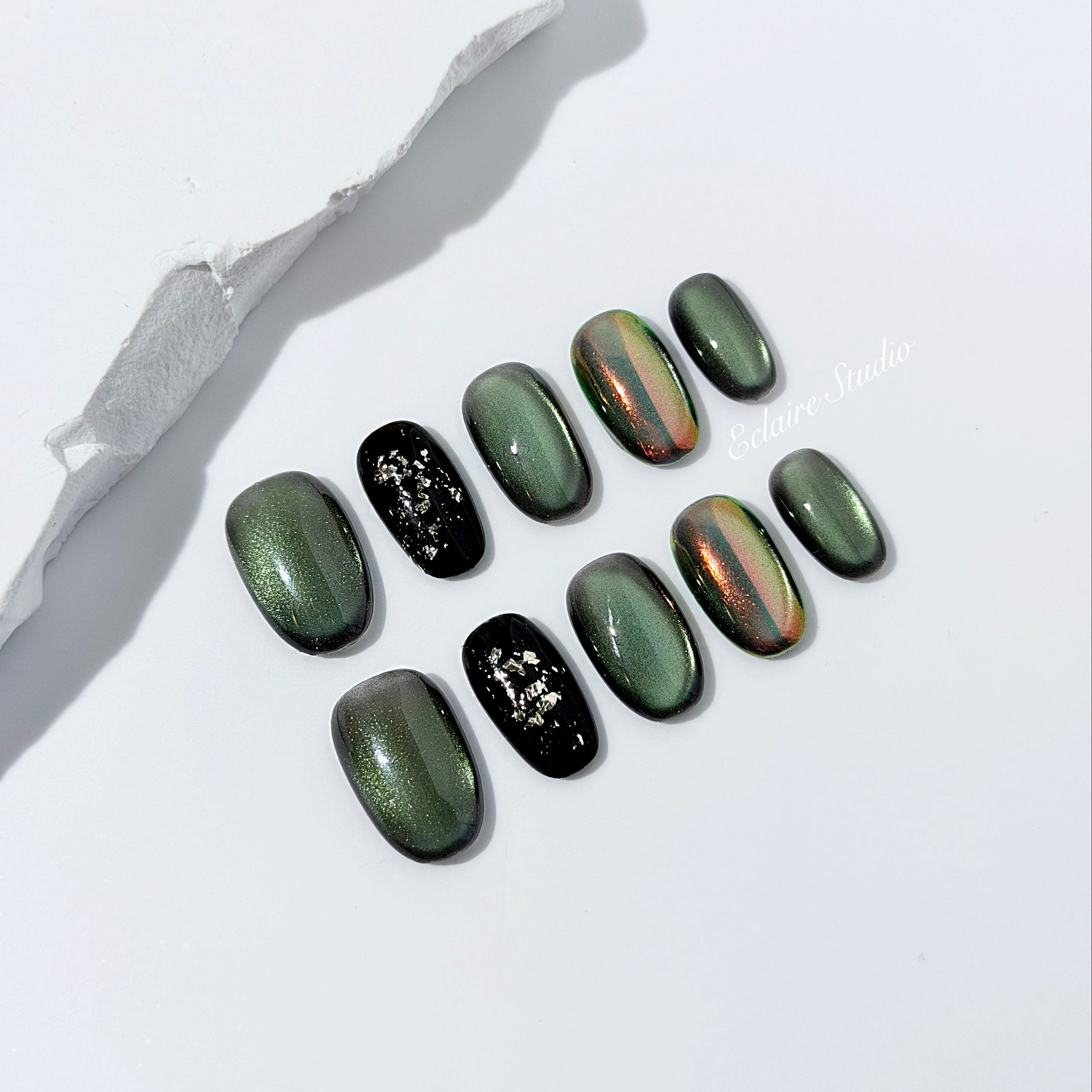 Short round press-on nails in shifting evergreen cat-eye shimmer with black and gold foil accent, displayed in a clear Éclaire Studio case.