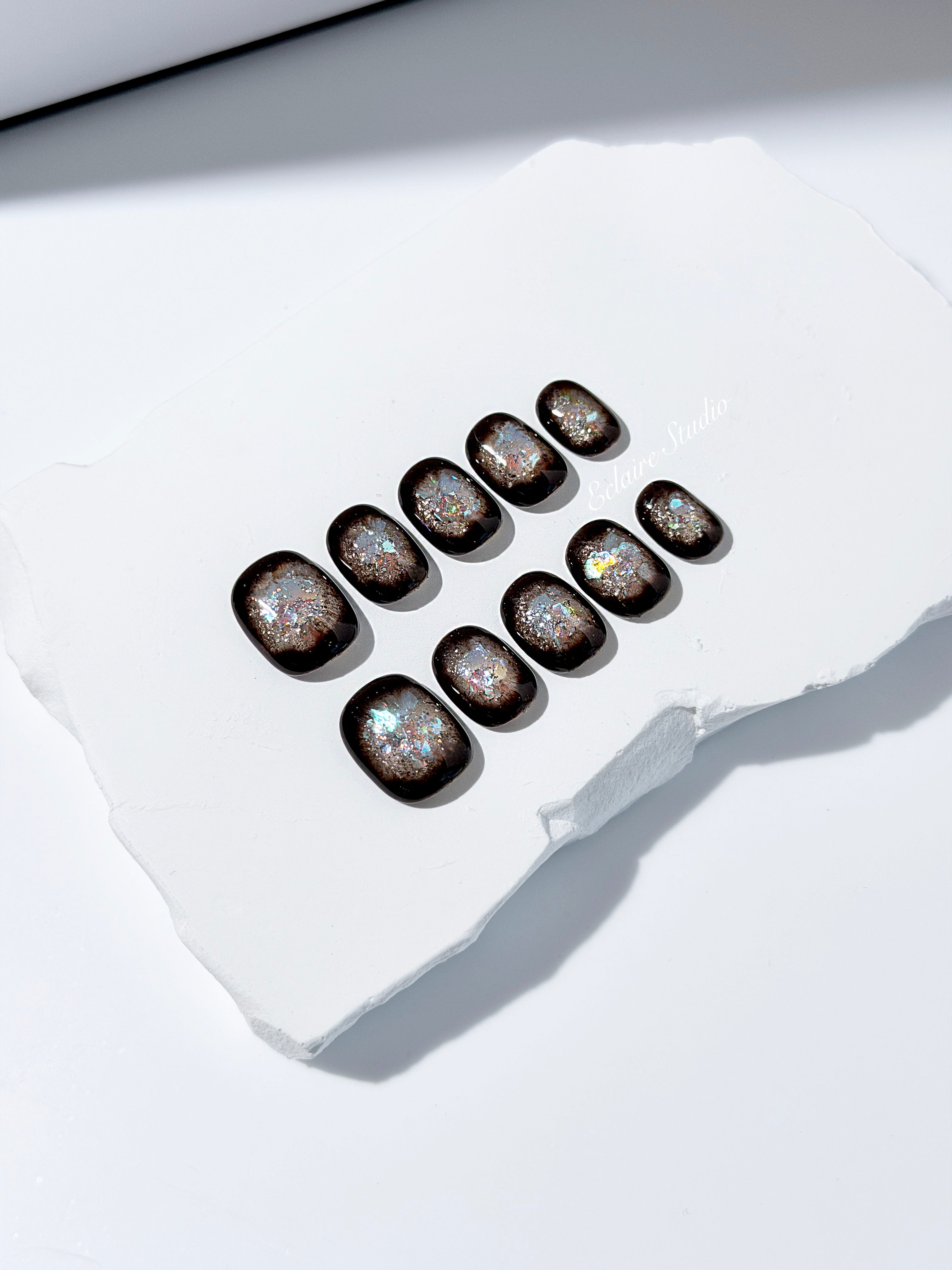 Short squoval press-on nails with dark brown base and iridescent frosted shimmer center, styled on white stone background.