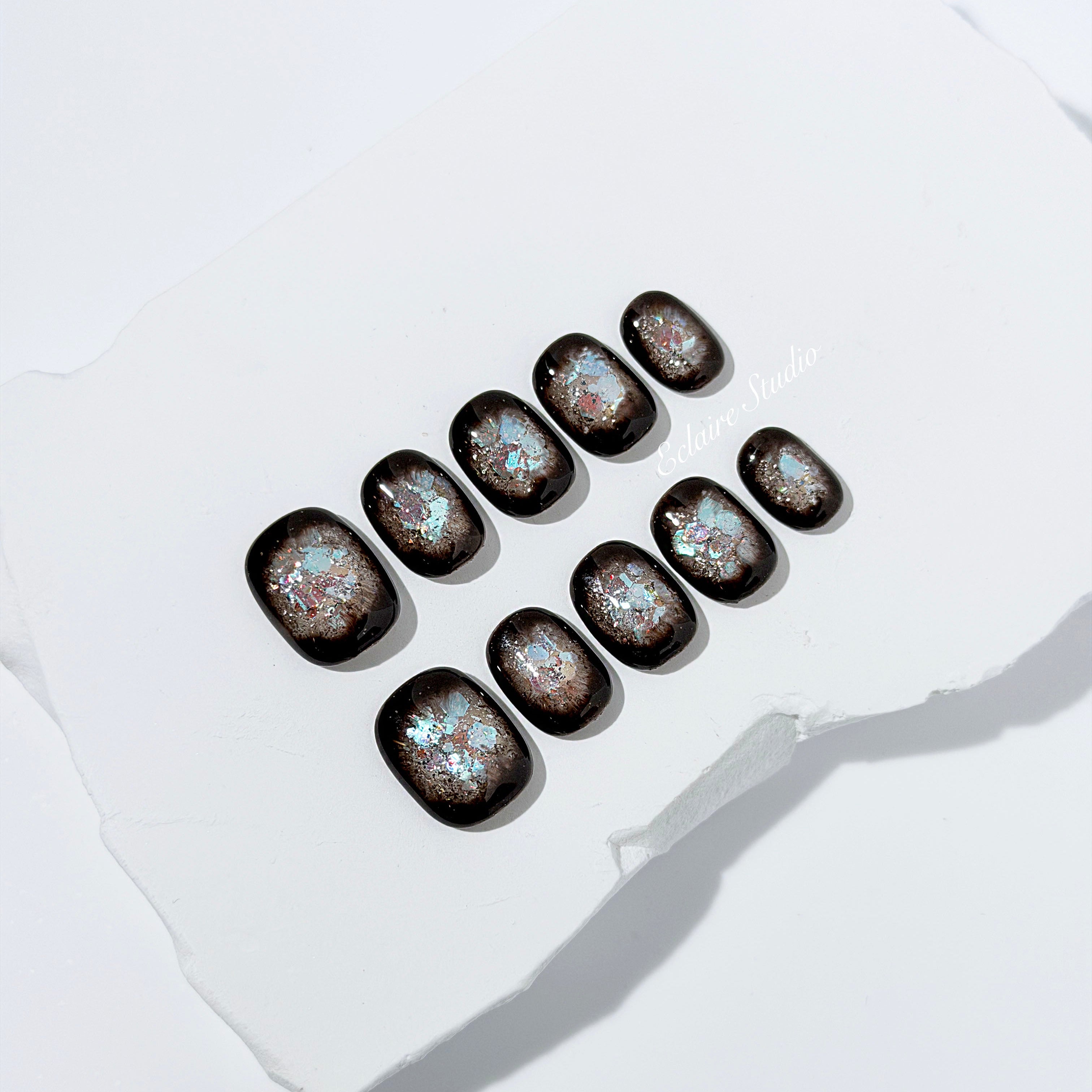 Short squoval press-on nails with dark brown base and iridescent frosted shimmer center, styled on white stone background.