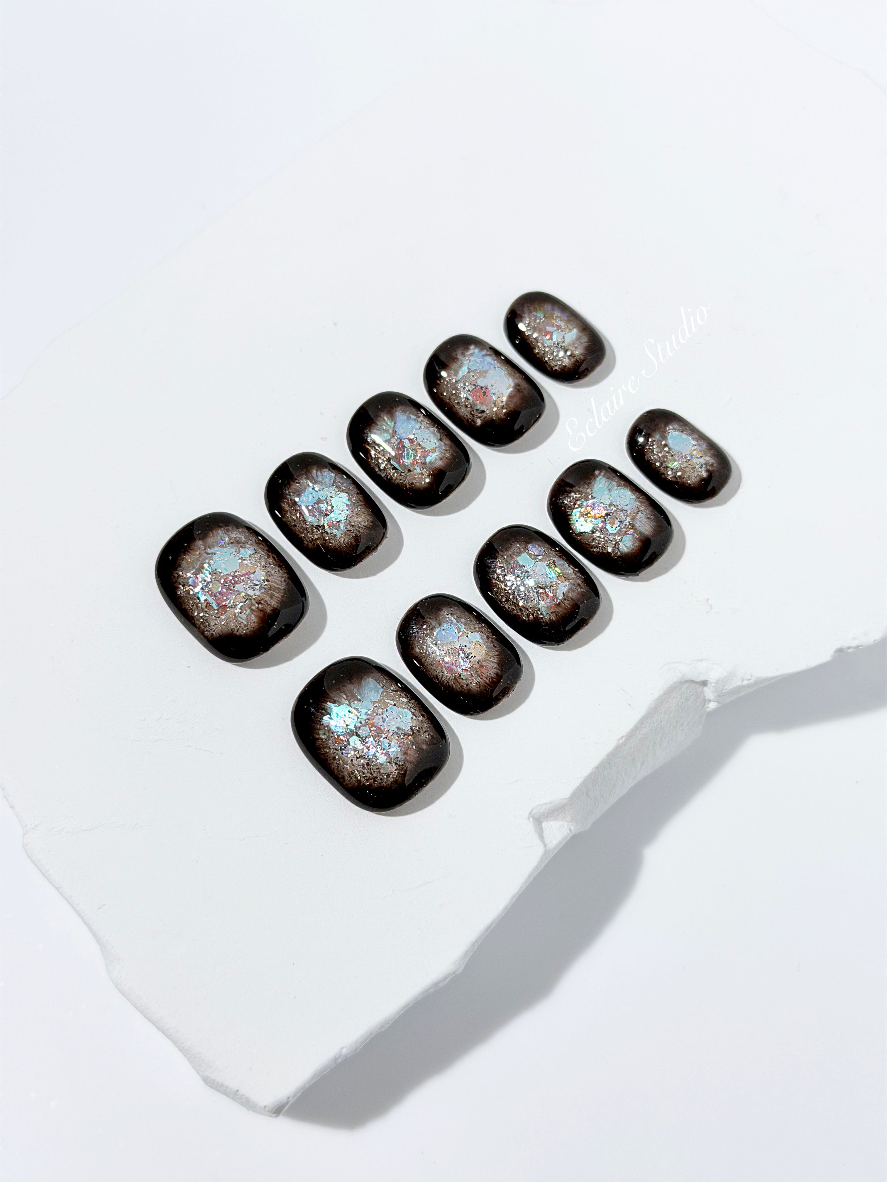 Short squoval press-on nails with dark brown base and iridescent frosted shimmer center, styled on white stone background.