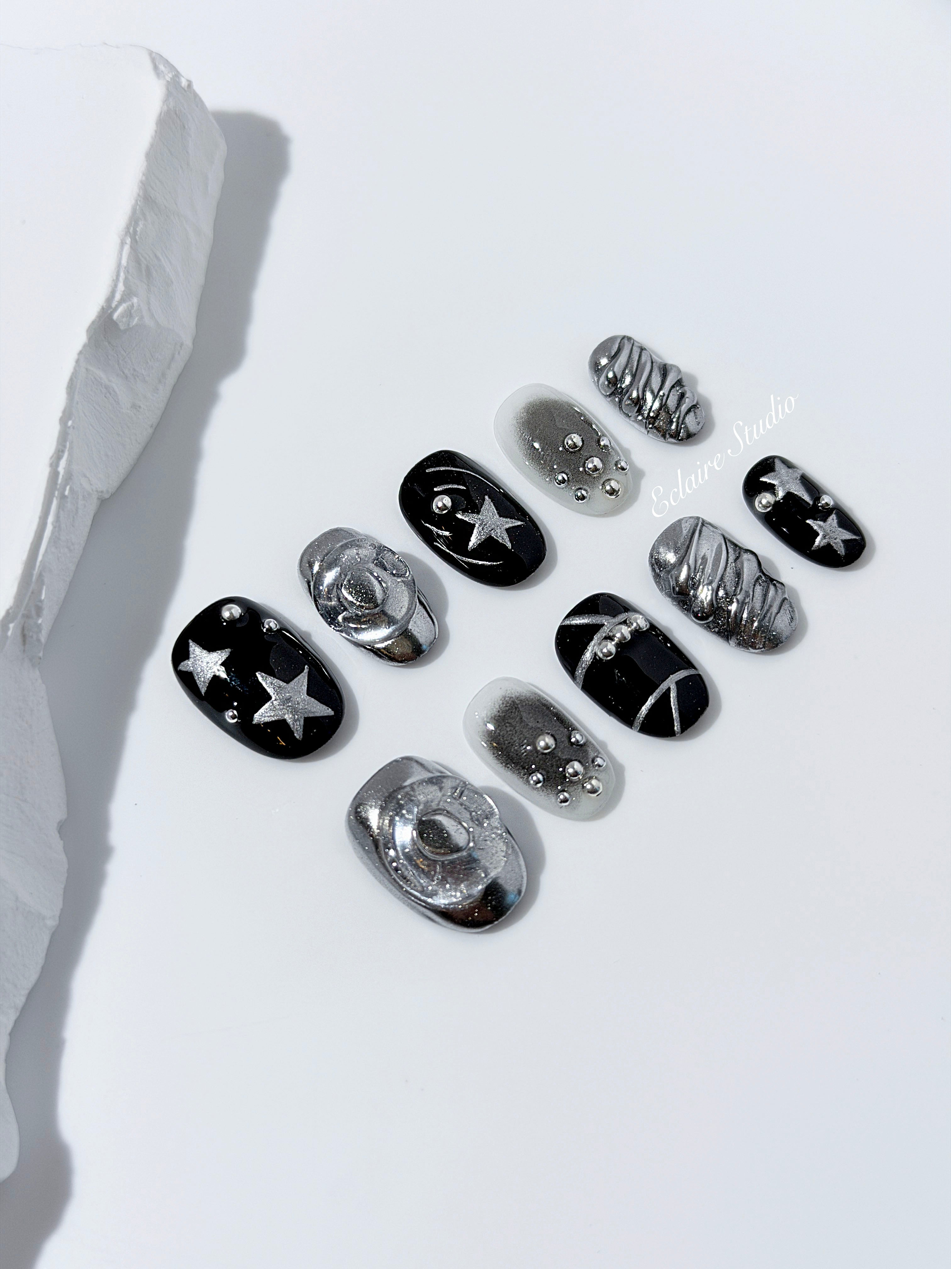 Black and silver press-on nails with 3D chrome swirl accents, star details, and shimmer gradient in a bold winter aesthetic.