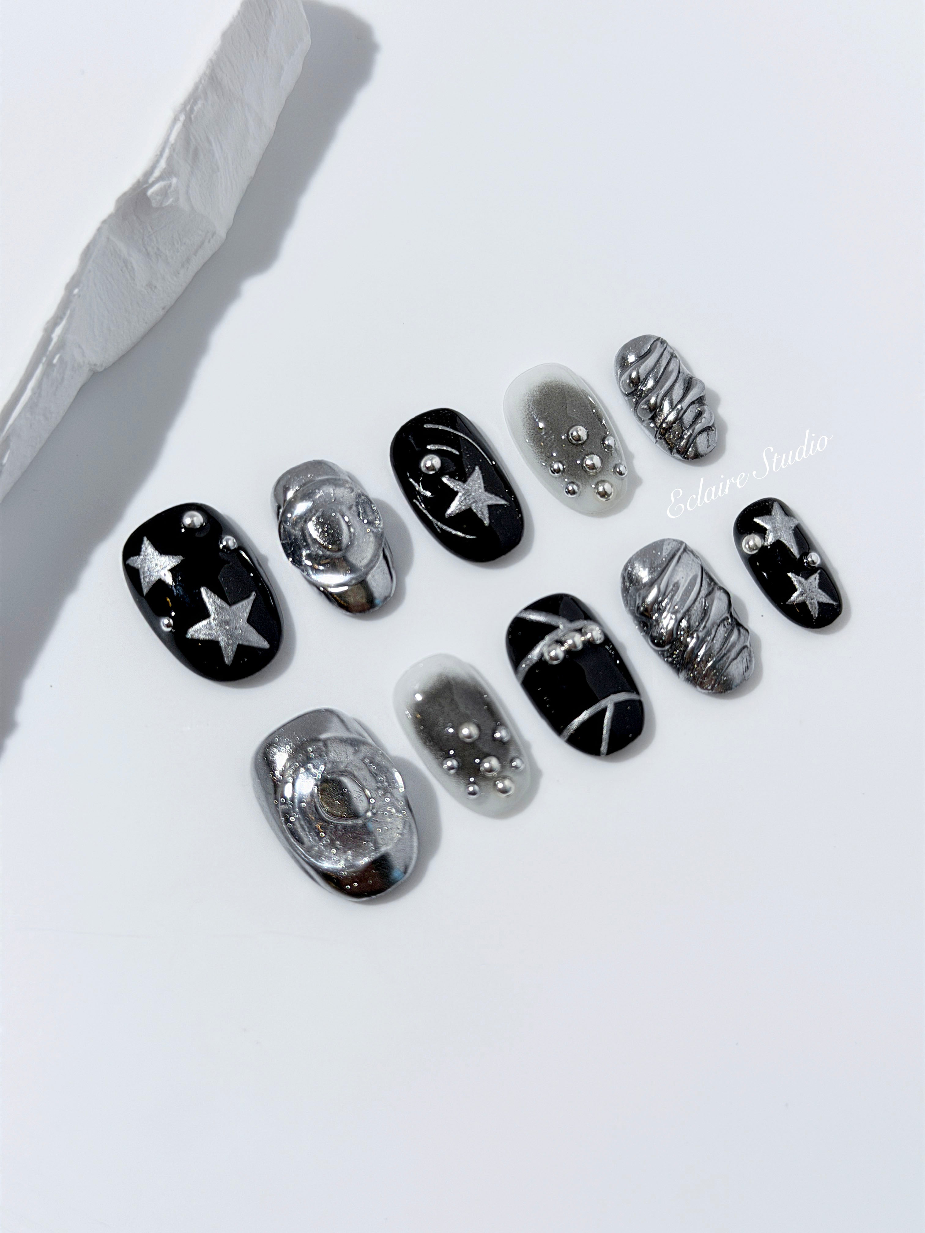 Black and silver press-on nails with 3D chrome swirl accents, star details, and shimmer gradient in a bold winter aesthetic.