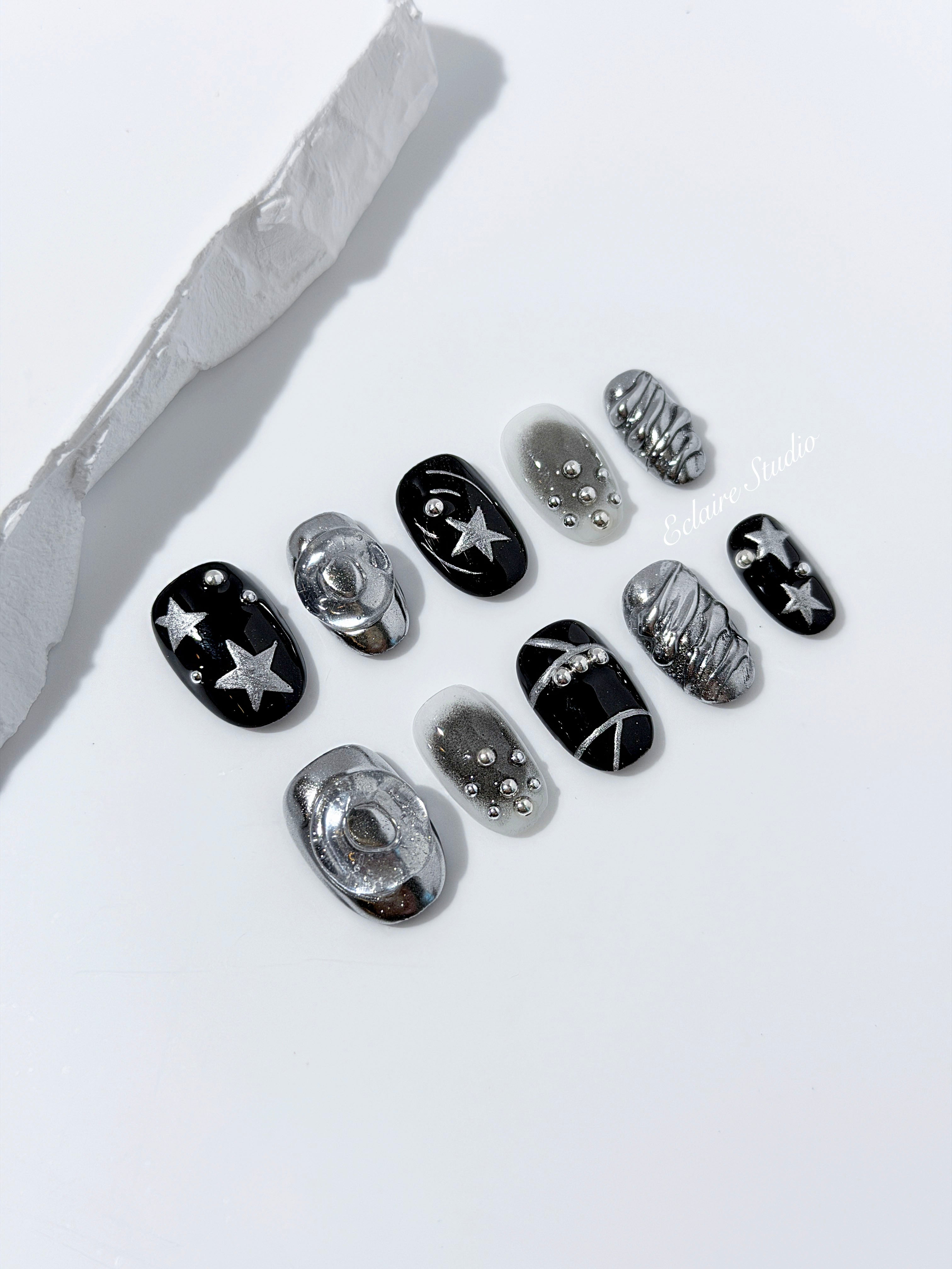 Black and silver press-on nails with 3D chrome swirl accents, star details, and shimmer gradient in a bold winter aesthetic.