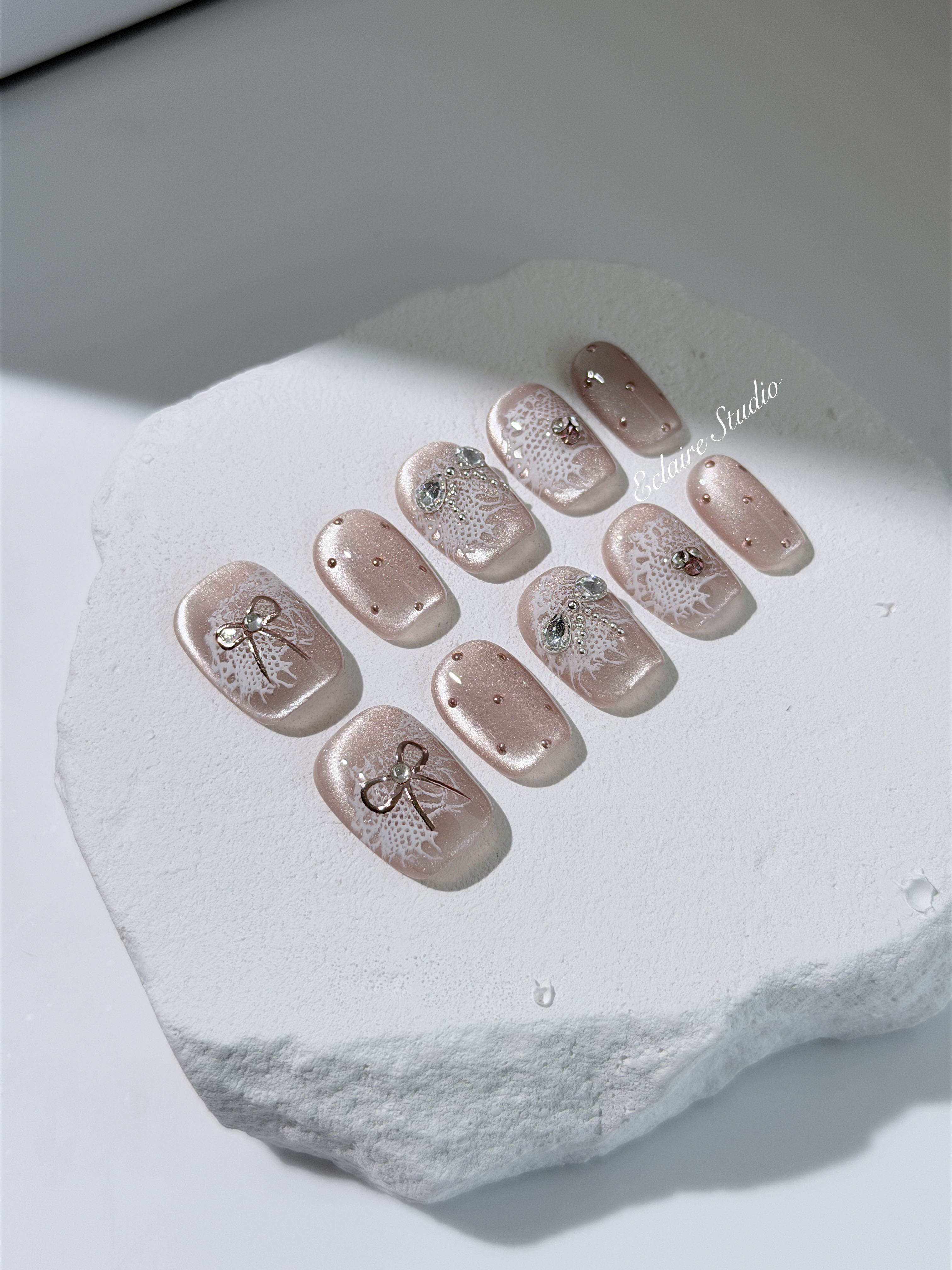 Short coffin pink nude cat-eye press-on nails with lace texture and pearl bow accents in a soft romantic winter style.