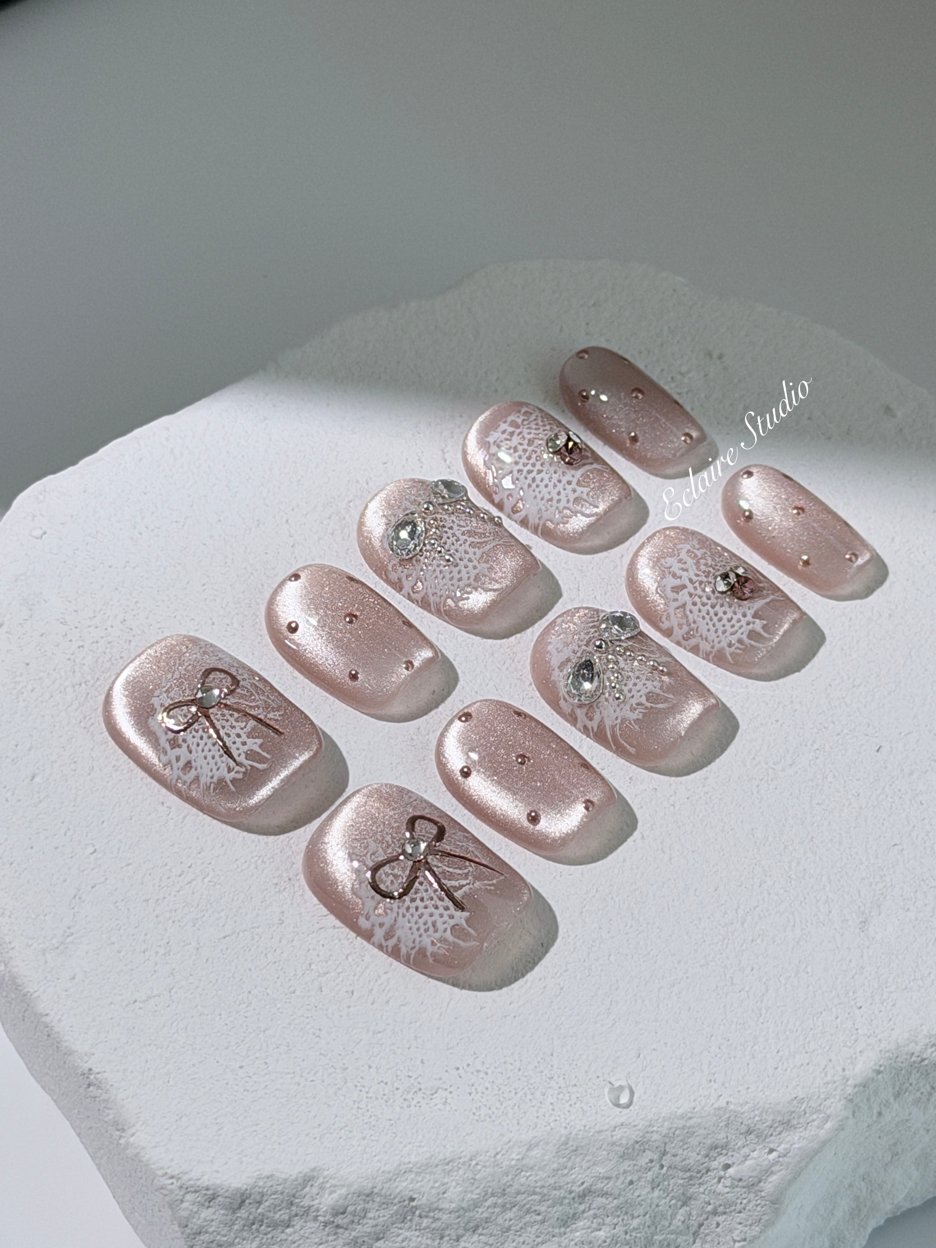 Short coffin pink nude cat-eye press-on nails with lace texture and pearl bow accents in a soft romantic winter style.