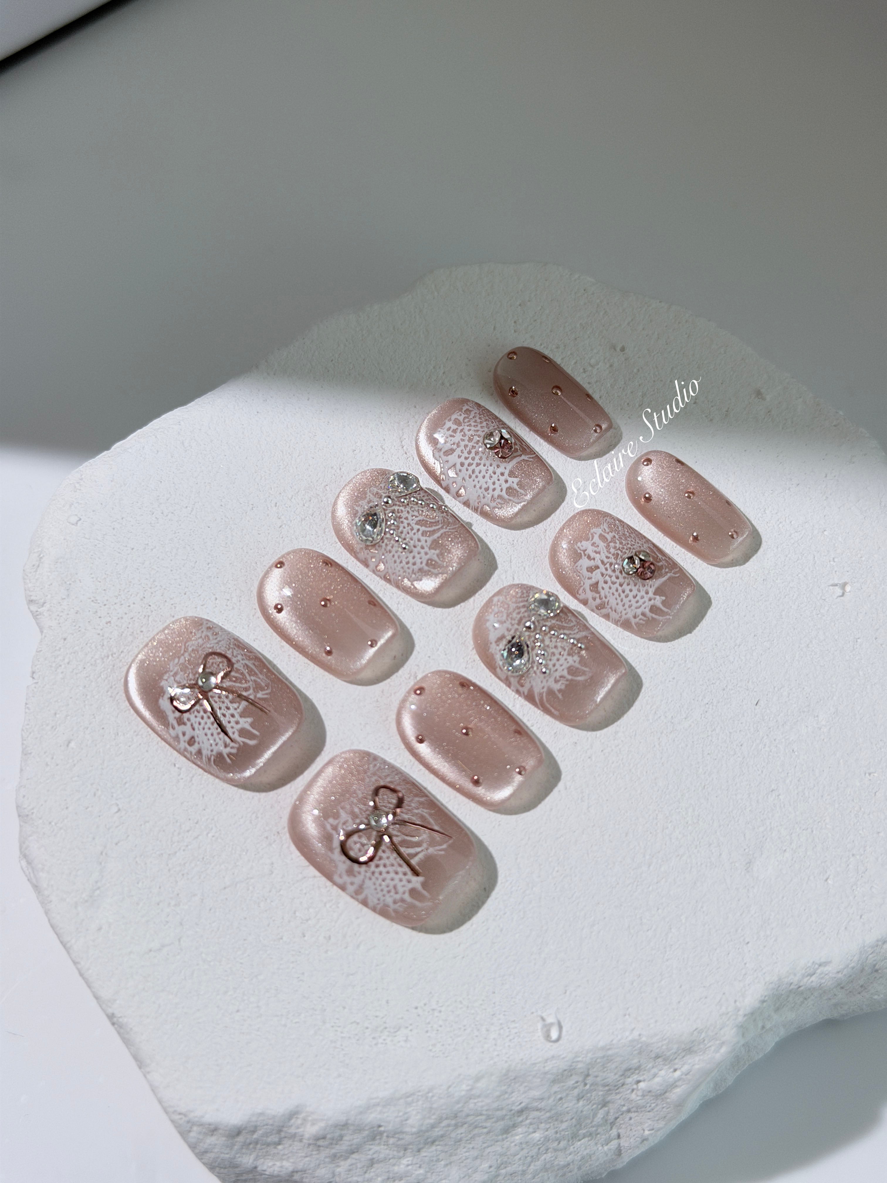 Short coffin pink nude cat-eye press-on nails with lace texture and pearl bow accents in a soft romantic winter style.
