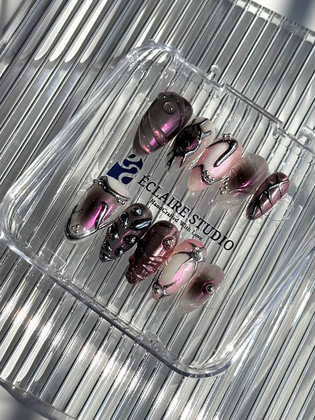 Set of long, almond-shaped press-on nails with a pink-to-purple multichrome base, accented by sculpted chrome designs, metallic textures, and small silver studs, displayed on a ribbed reflective surface under natural light.