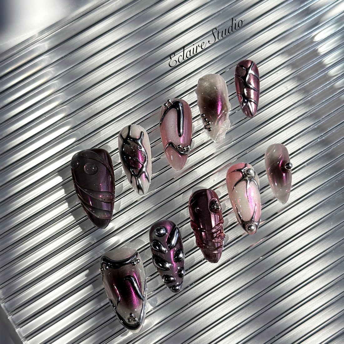 Set of long, almond-shaped press-on nails with a pink-to-purple multichrome base, accented by sculpted chrome designs, metallic textures, and small silver studs, displayed on a ribbed reflective surface under natural light.