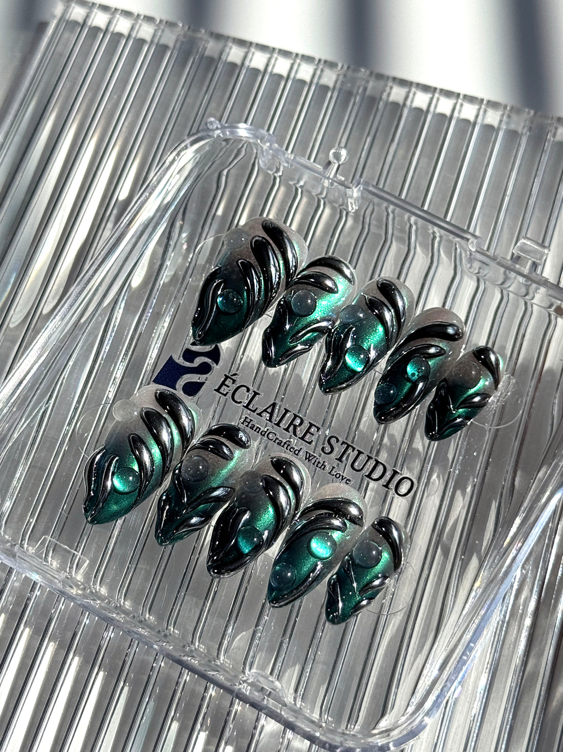 Set of long almond-shaped press-on nails featuring a smoky green gradient base with metallic emerald green shimmer, accented by bold black chrome sculpted designs and glossy 3D details, displayed on a ribbed reflective surface under bright light.