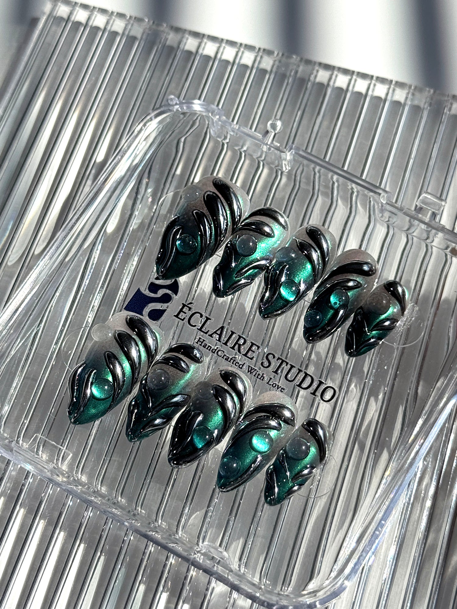 Set of long almond-shaped press-on nails featuring a smoky green gradient base with metallic emerald green shimmer, accented by bold black chrome sculpted designs and glossy 3D details, displayed on a ribbed reflective surface under bright light.