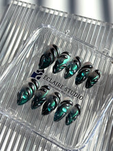 Set of long almond-shaped press-on nails featuring a smoky green gradient base with metallic emerald green shimmer, accented by bold black chrome sculpted designs and glossy 3D details, displayed on a ribbed reflective surface under bright light.