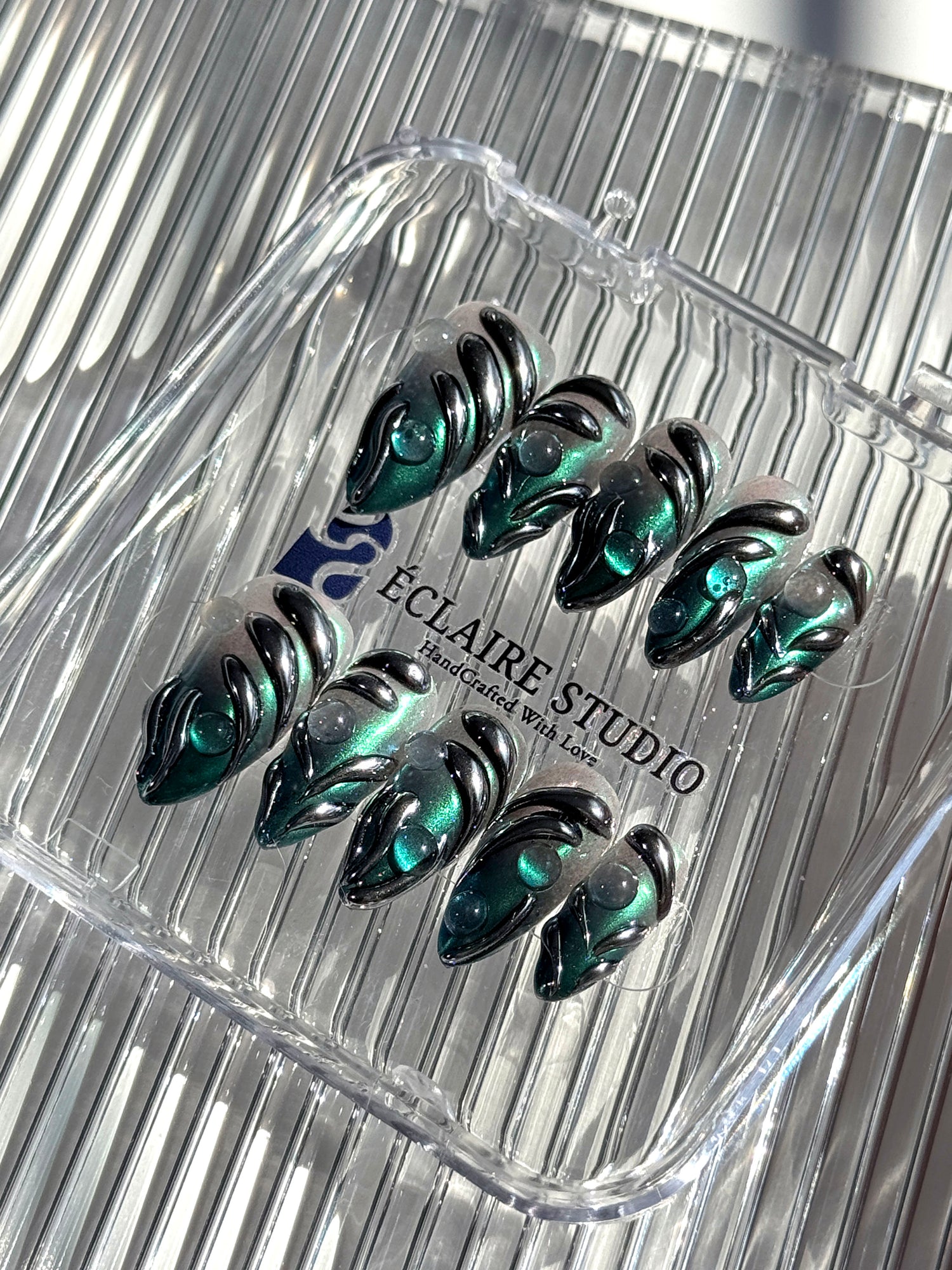 Set of long almond-shaped press-on nails featuring a smoky green gradient base with metallic emerald green shimmer, accented by bold black chrome sculpted designs and glossy 3D details, displayed on a ribbed reflective surface under bright light.