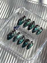 Set of long almond-shaped press-on nails featuring a smoky green gradient base with metallic emerald green shimmer, accented by bold black chrome sculpted designs and glossy 3D details, displayed on a ribbed reflective surface under bright light.