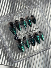 Set of long almond-shaped press-on nails featuring a smoky green gradient base with metallic emerald green shimmer, accented by bold black chrome sculpted designs and glossy 3D details, displayed on a ribbed reflective surface under bright light.