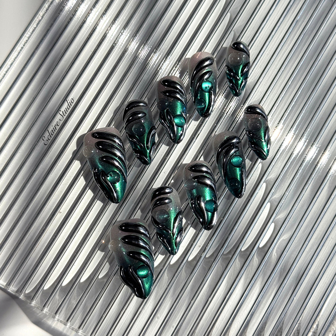Set of long almond-shaped press-on nails featuring a smoky green gradient base with metallic emerald green shimmer, accented by bold black chrome sculpted designs and glossy 3D details, displayed on a ribbed reflective surface under bright light.