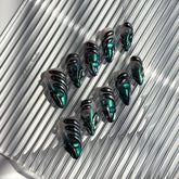 Set of long almond-shaped press-on nails featuring a smoky green gradient base with metallic emerald green shimmer, accented by bold black chrome sculpted designs and glossy 3D details, displayed on a ribbed reflective surface under bright light.