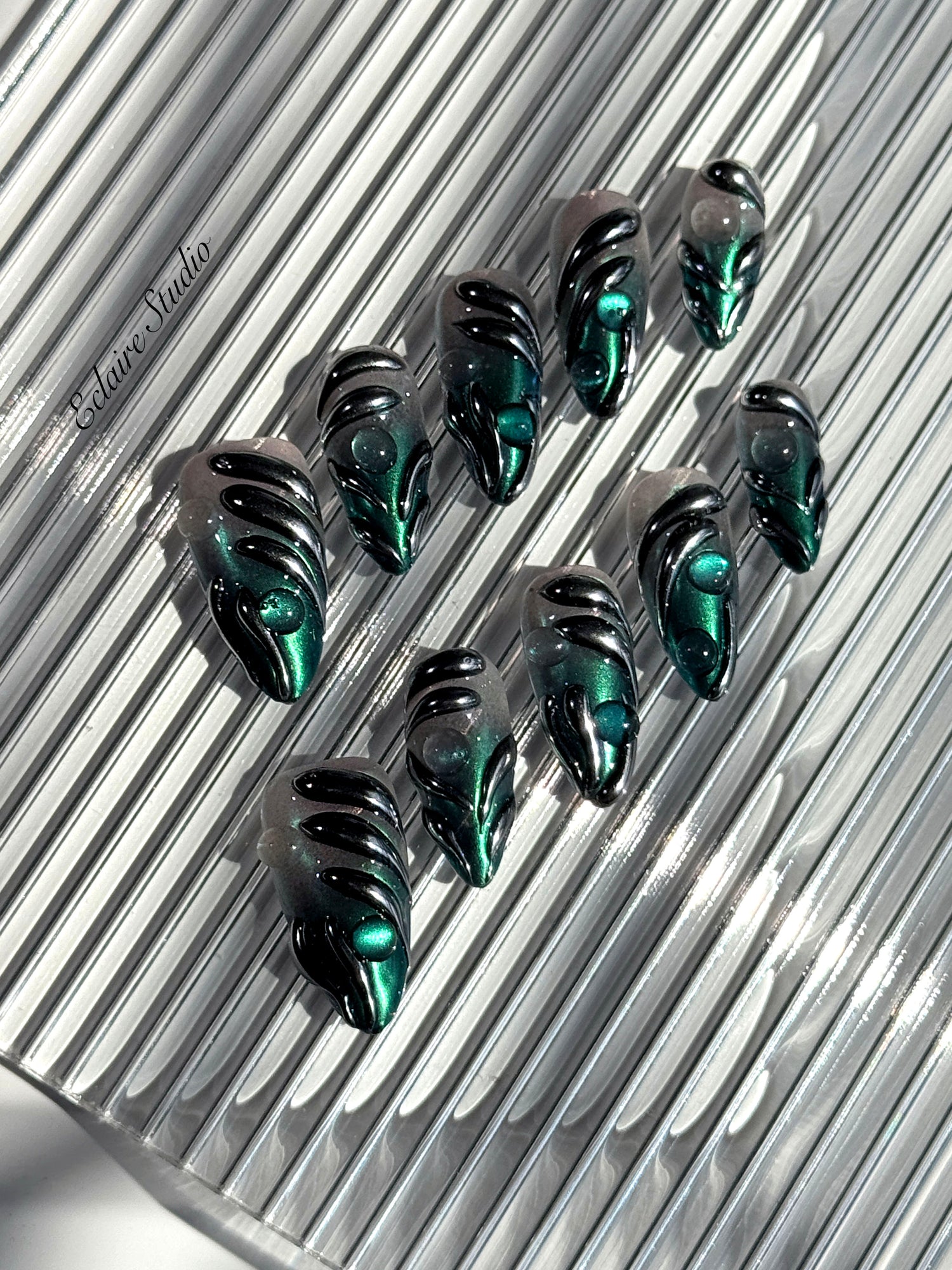 Set of long almond-shaped press-on nails featuring a smoky green gradient base with metallic emerald green shimmer, accented by bold black chrome sculpted designs and glossy 3D details, displayed on a ribbed reflective surface under bright light.