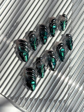 Set of long almond-shaped press-on nails featuring a smoky green gradient base with metallic emerald green shimmer, accented by bold black chrome sculpted designs and glossy 3D details, displayed on a ribbed reflective surface under bright light.