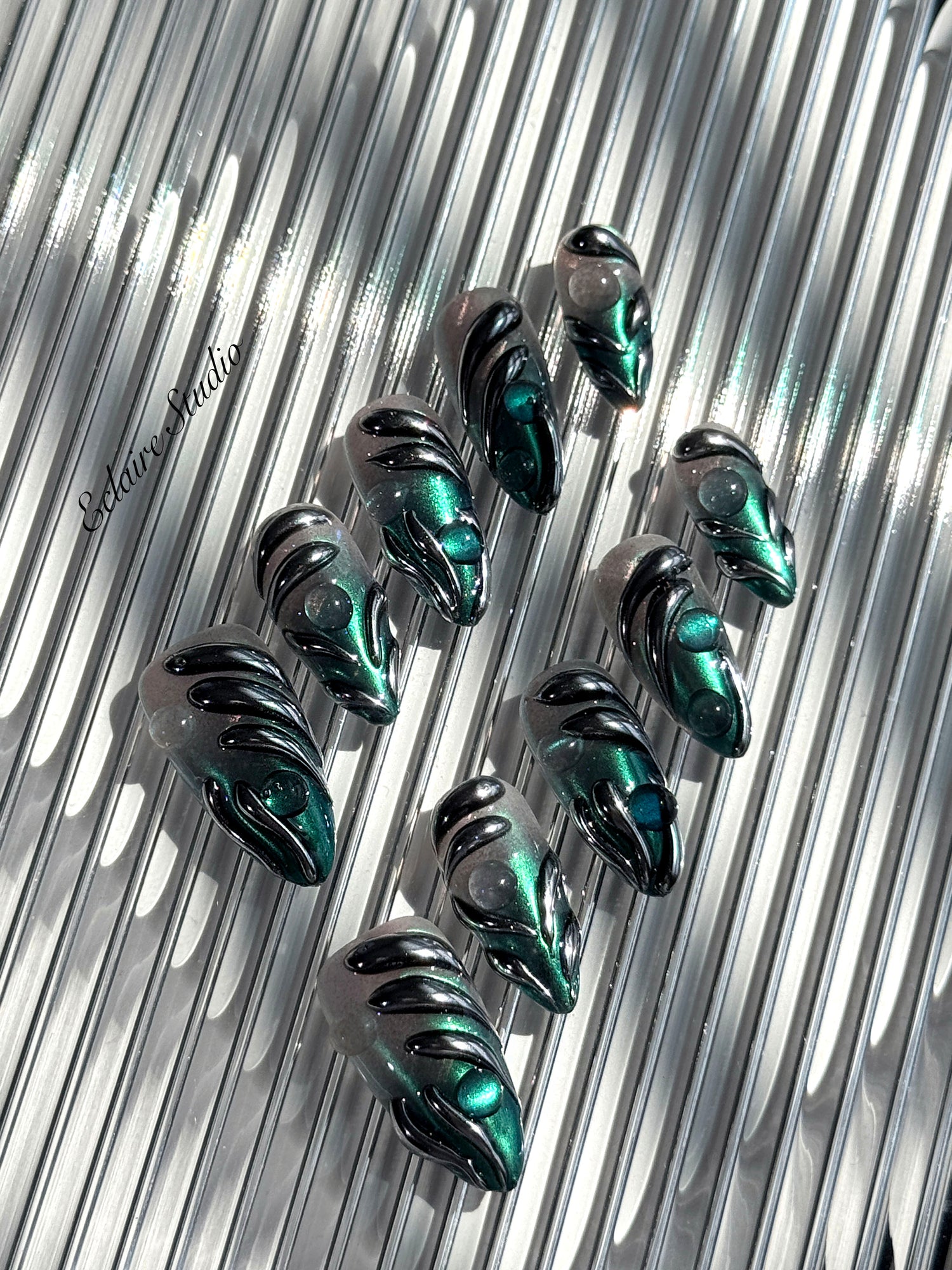 Set of long almond-shaped press-on nails featuring a smoky green gradient base with metallic emerald green shimmer, accented by bold black chrome sculpted designs and glossy 3D details, displayed on a ribbed reflective surface under bright light.