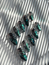 Set of long almond-shaped press-on nails featuring a smoky green gradient base with metallic emerald green shimmer, accented by bold black chrome sculpted designs and glossy 3D details, displayed on a ribbed reflective surface under bright light.