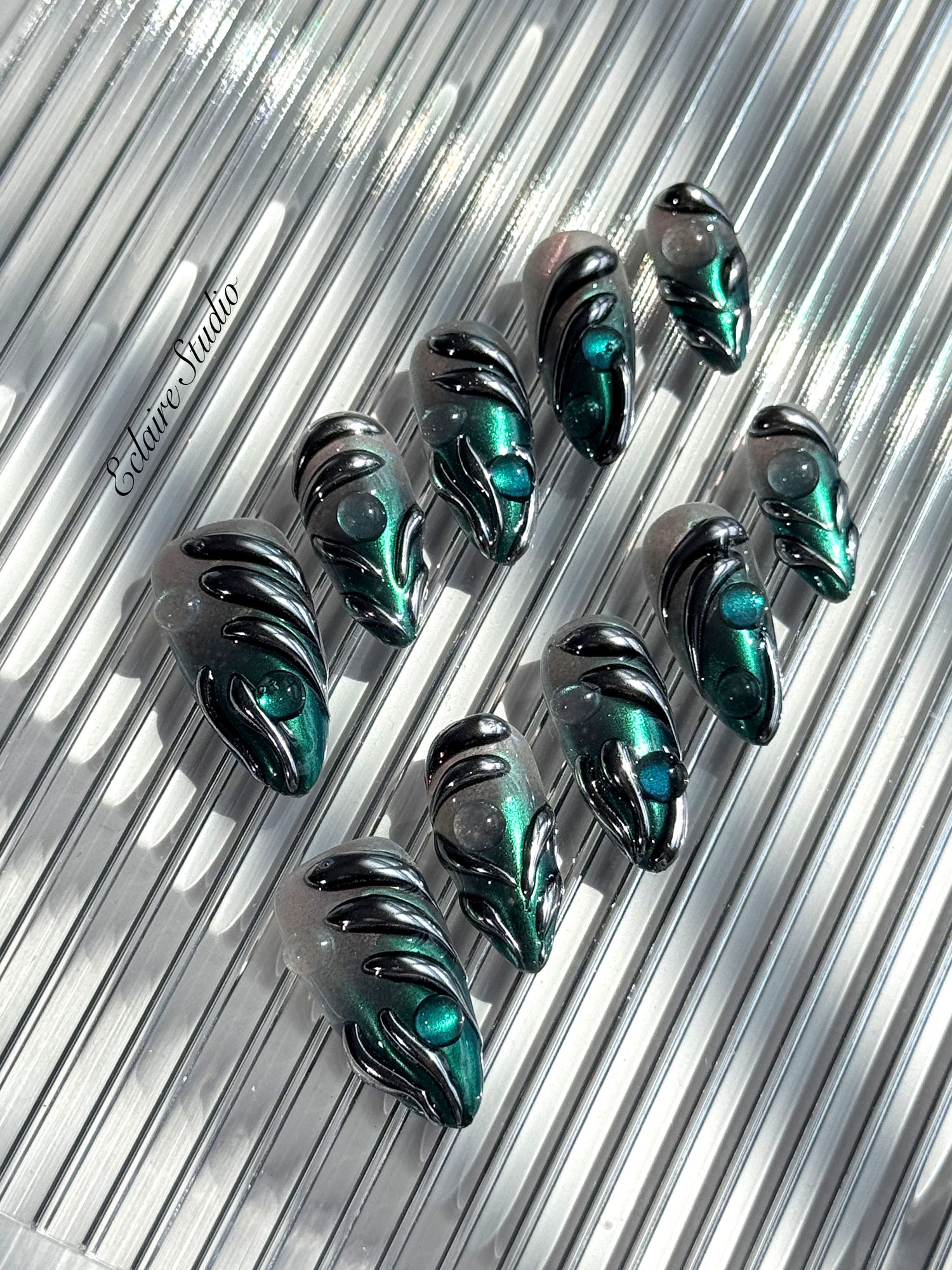 Set of long almond-shaped press-on nails featuring a smoky green gradient base with metallic emerald green shimmer, accented by bold black chrome sculpted designs and glossy 3D details, displayed on a ribbed reflective surface under bright light.