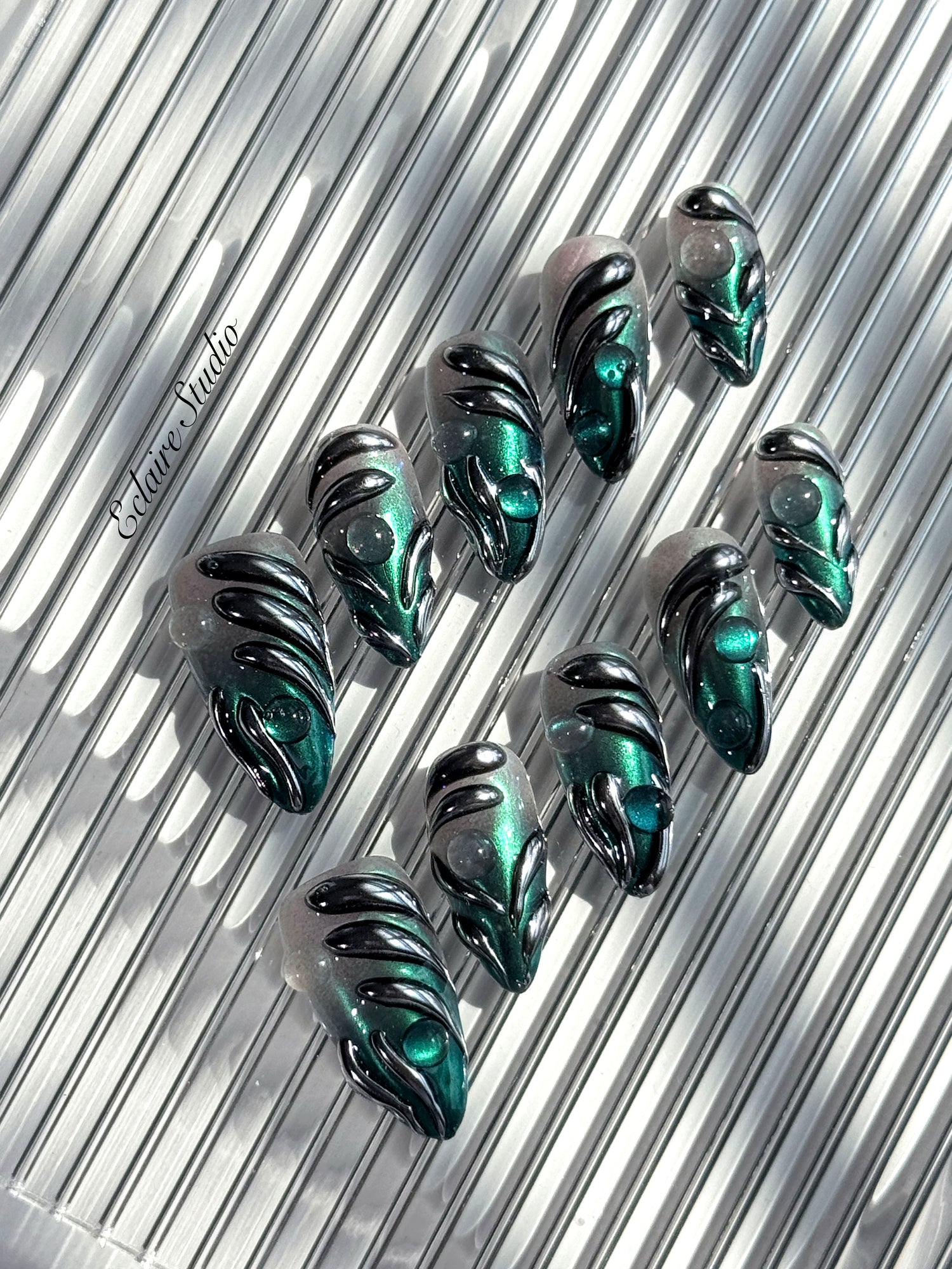 Set of long almond-shaped press-on nails featuring a smoky green gradient base with metallic emerald green shimmer, accented by bold black chrome sculpted designs and glossy 3D details, displayed on a ribbed reflective surface under bright light.