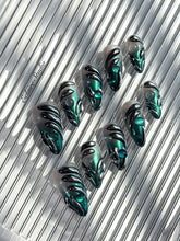 Set of long almond-shaped press-on nails featuring a smoky green gradient base with metallic emerald green shimmer, accented by bold black chrome sculpted designs and glossy 3D details, displayed on a ribbed reflective surface under bright light.