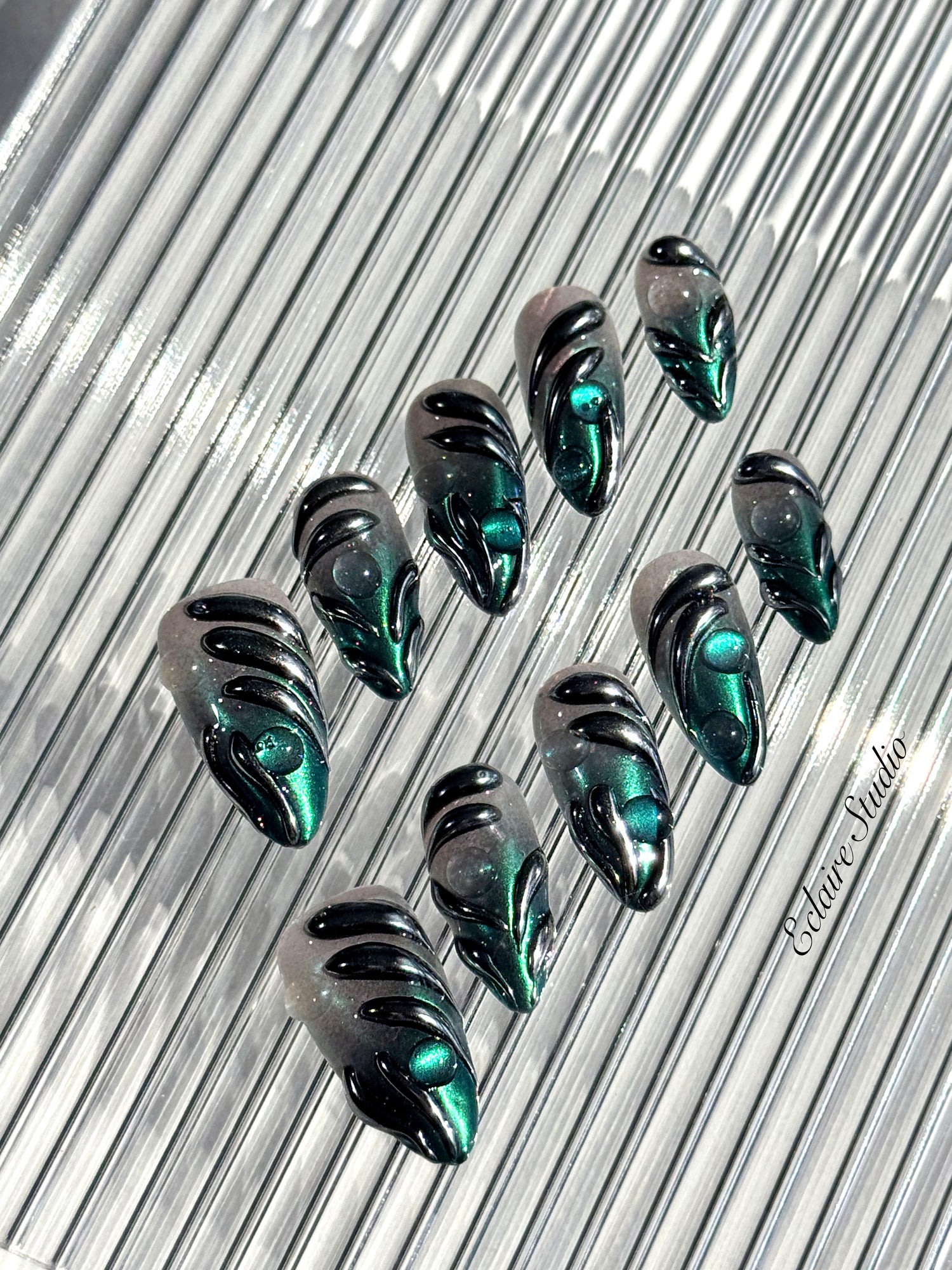Set of long almond-shaped press-on nails featuring a smoky green gradient base with metallic emerald green shimmer, accented by bold black chrome sculpted designs and glossy 3D details, displayed on a ribbed reflective surface under bright light.