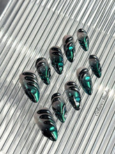 Set of long almond-shaped press-on nails featuring a smoky green gradient base with metallic emerald green shimmer, accented by bold black chrome sculpted designs and glossy 3D details, displayed on a ribbed reflective surface under bright light.
