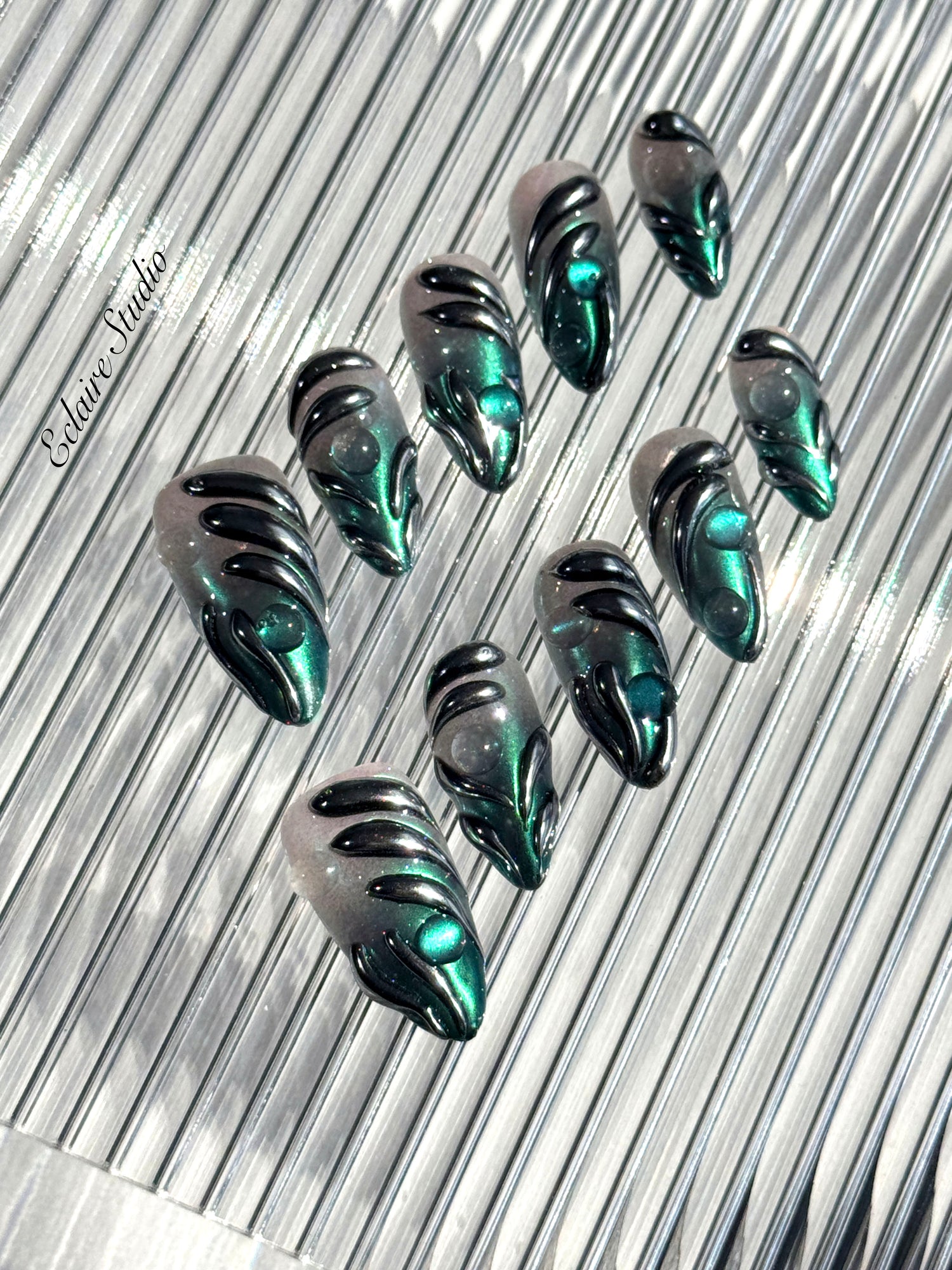 Set of long almond-shaped press-on nails featuring a smoky green gradient base with metallic emerald green shimmer, accented by bold black chrome sculpted designs and glossy 3D details, displayed on a ribbed reflective surface under bright light.