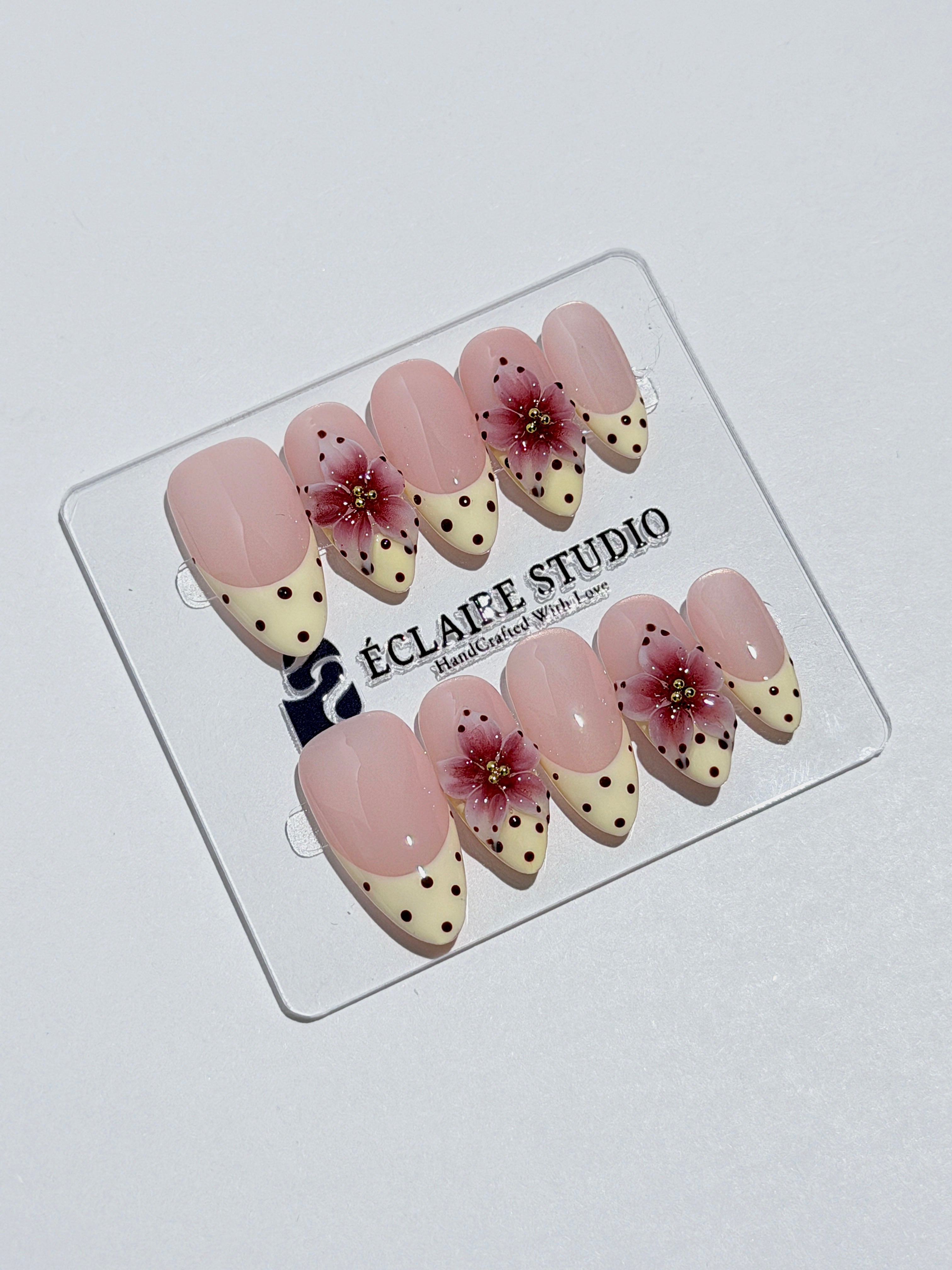 Luxury medium almond press-on nails featuring sheer pink bases with vintage cream French tips, burgundy polka dots, and hand-sculpted 3D burgundy flowers with gold bead centers.