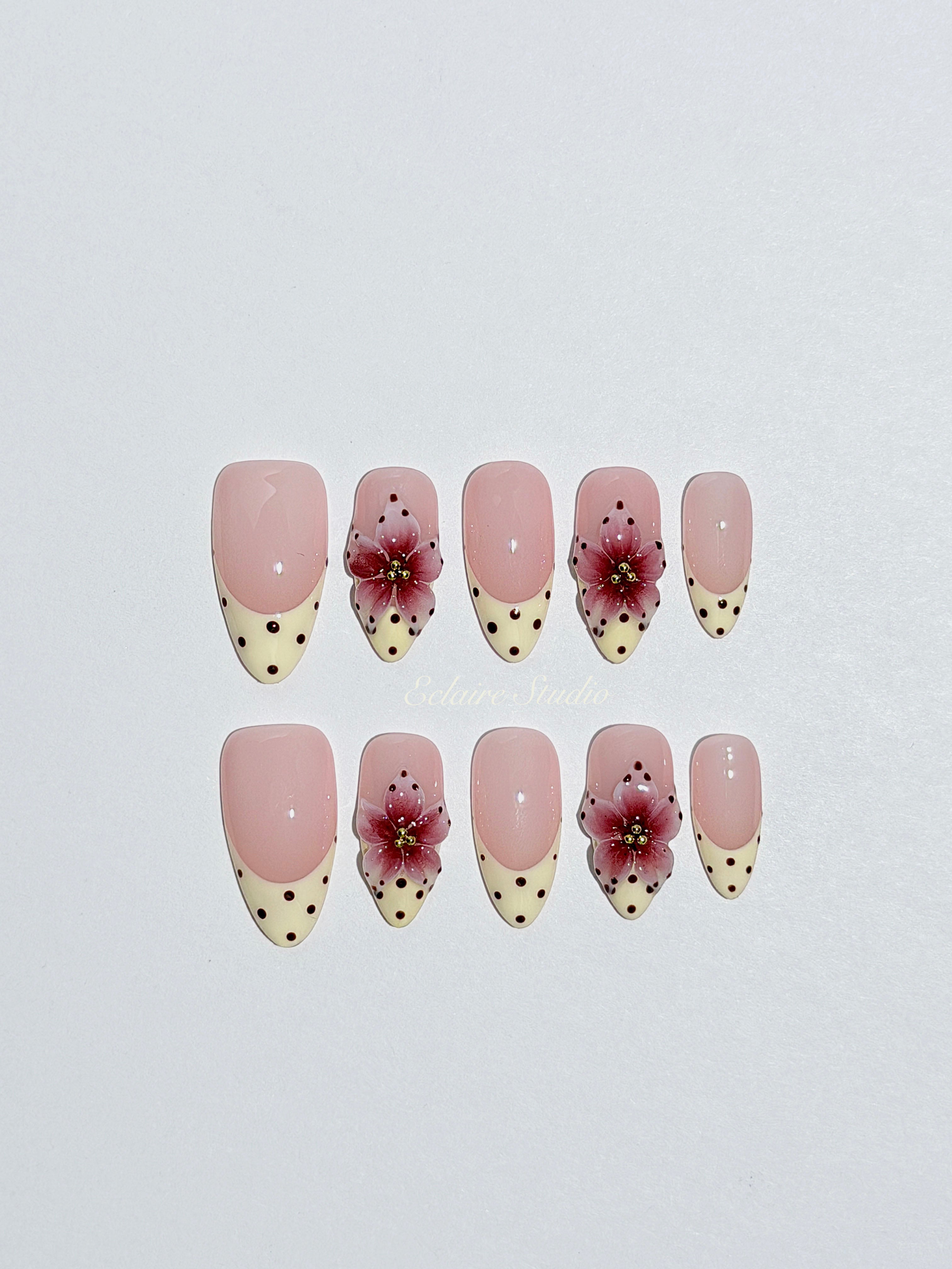 Luxury medium almond press-on nails featuring sheer pink bases with vintage cream French tips, burgundy polka dots, and hand-sculpted 3D burgundy flowers with gold bead centers.