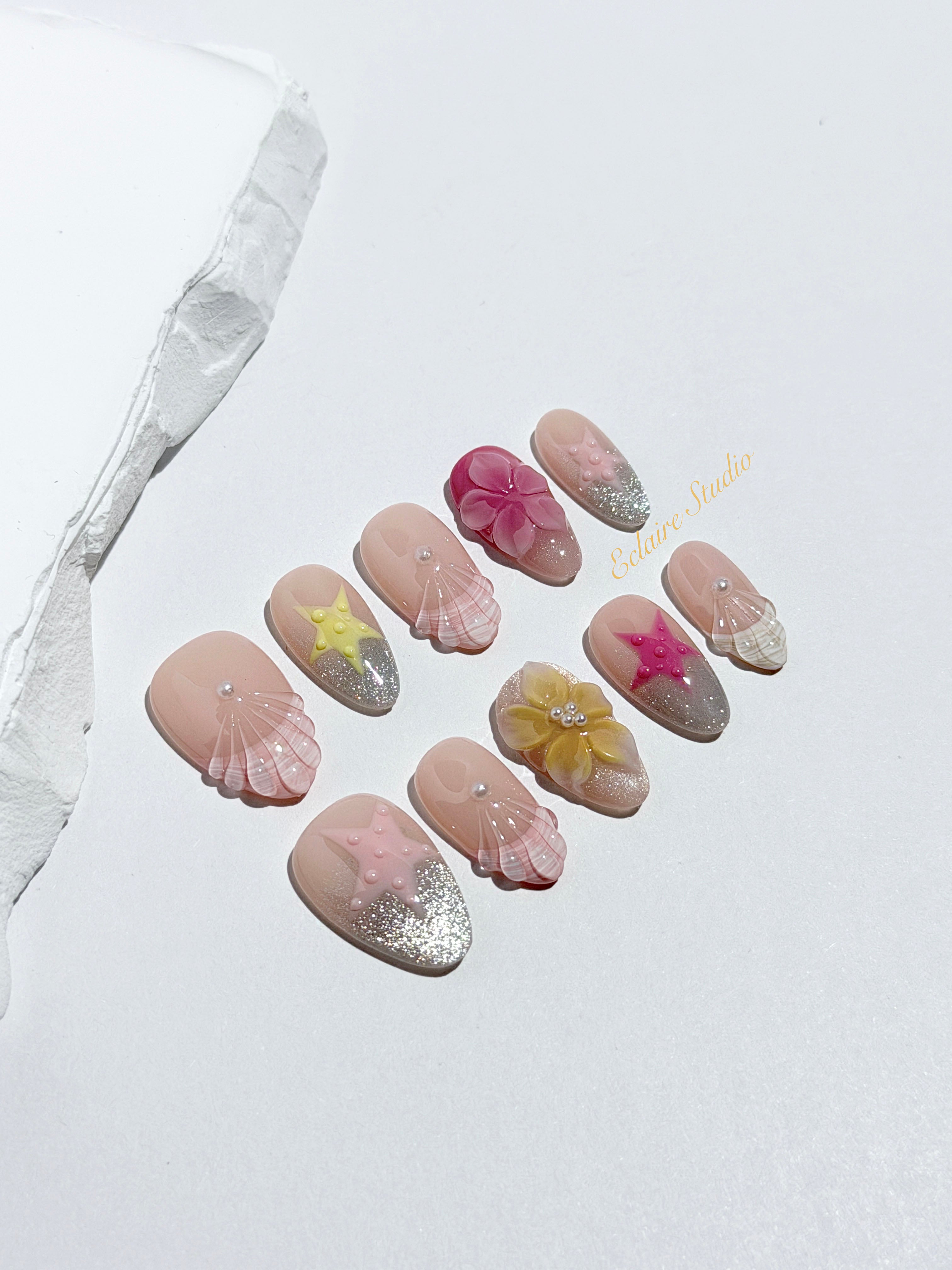 Eclaire Studio 3D Summer short almond press-on nails with sheer nude bases, sparkling silver cat-eye magnetic French tips, hand-painted colorful starfish, and pink 3D sculpted seashells.