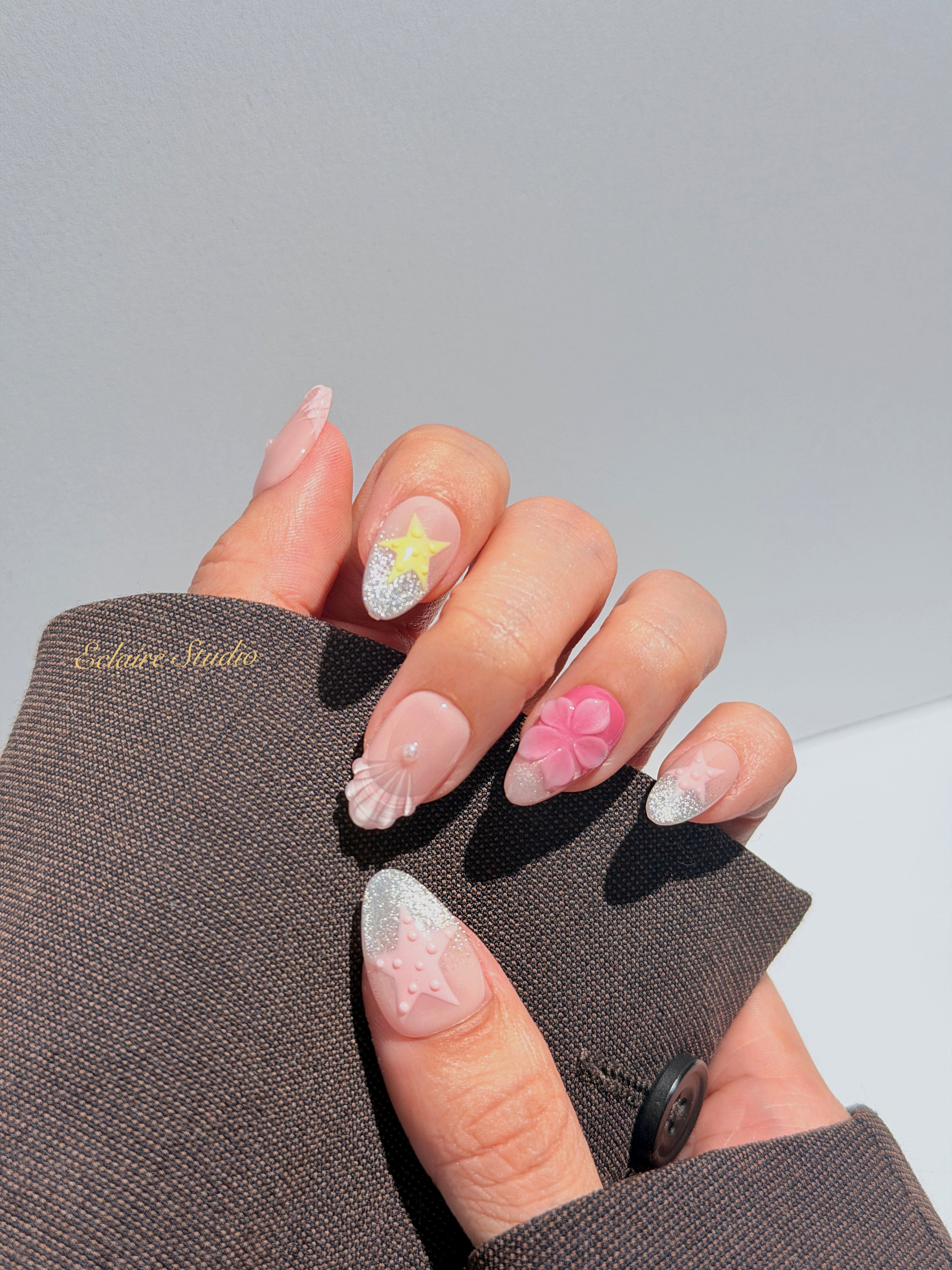 Eclaire Studio 3D Summer short almond press-on nails with sheer nude bases, sparkling silver cat-eye magnetic French tips, hand-painted colorful starfish, and pink 3D sculpted seashells.