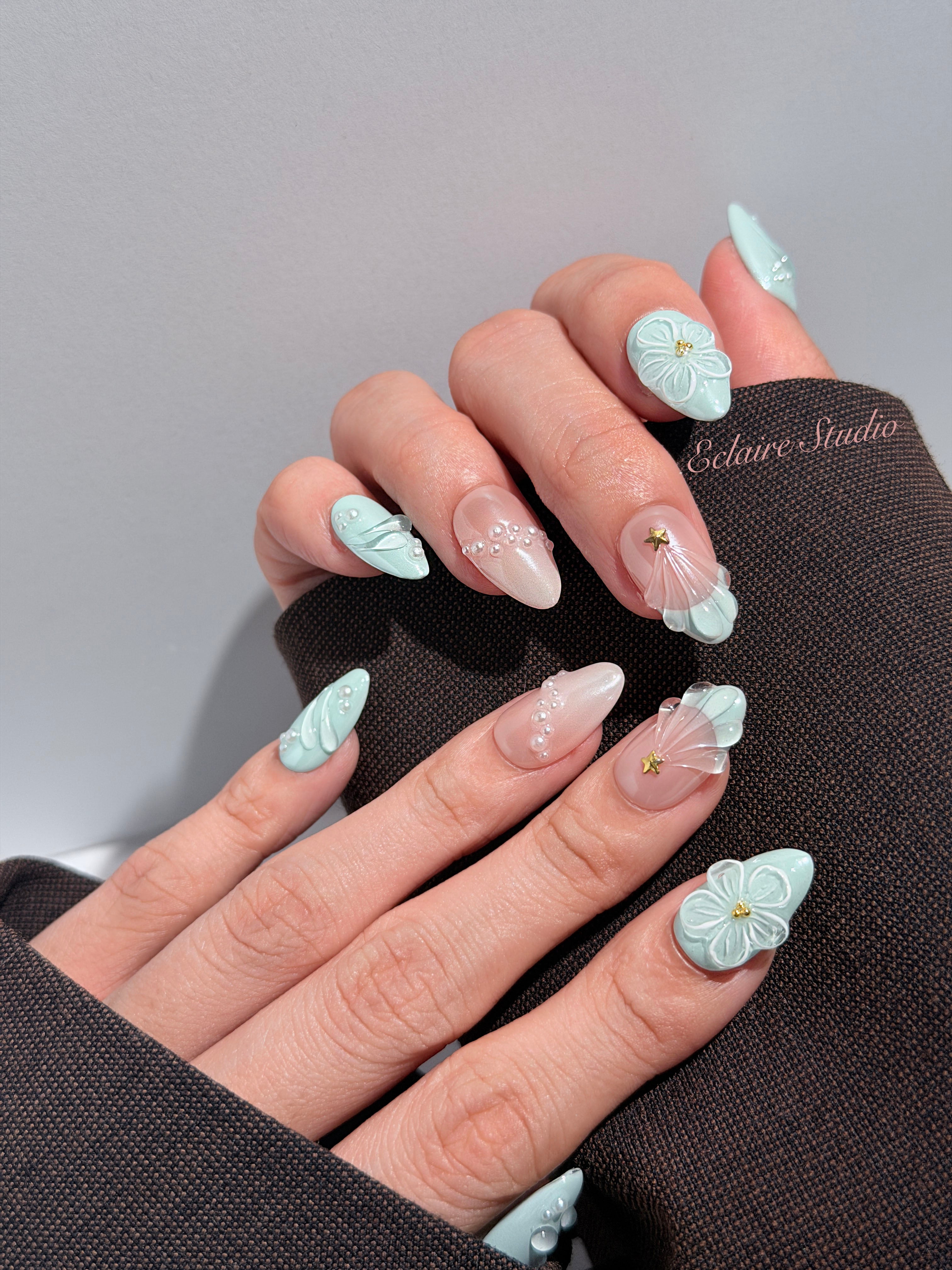 Eclaire Nail Studio 3D Summer short almond press-on nails featuring a light aqua glazed base and peachy-nude accent nails with 3D sculpted ocean waves, 3D flowers, and seashell tips with mini pearls and gold stars.
