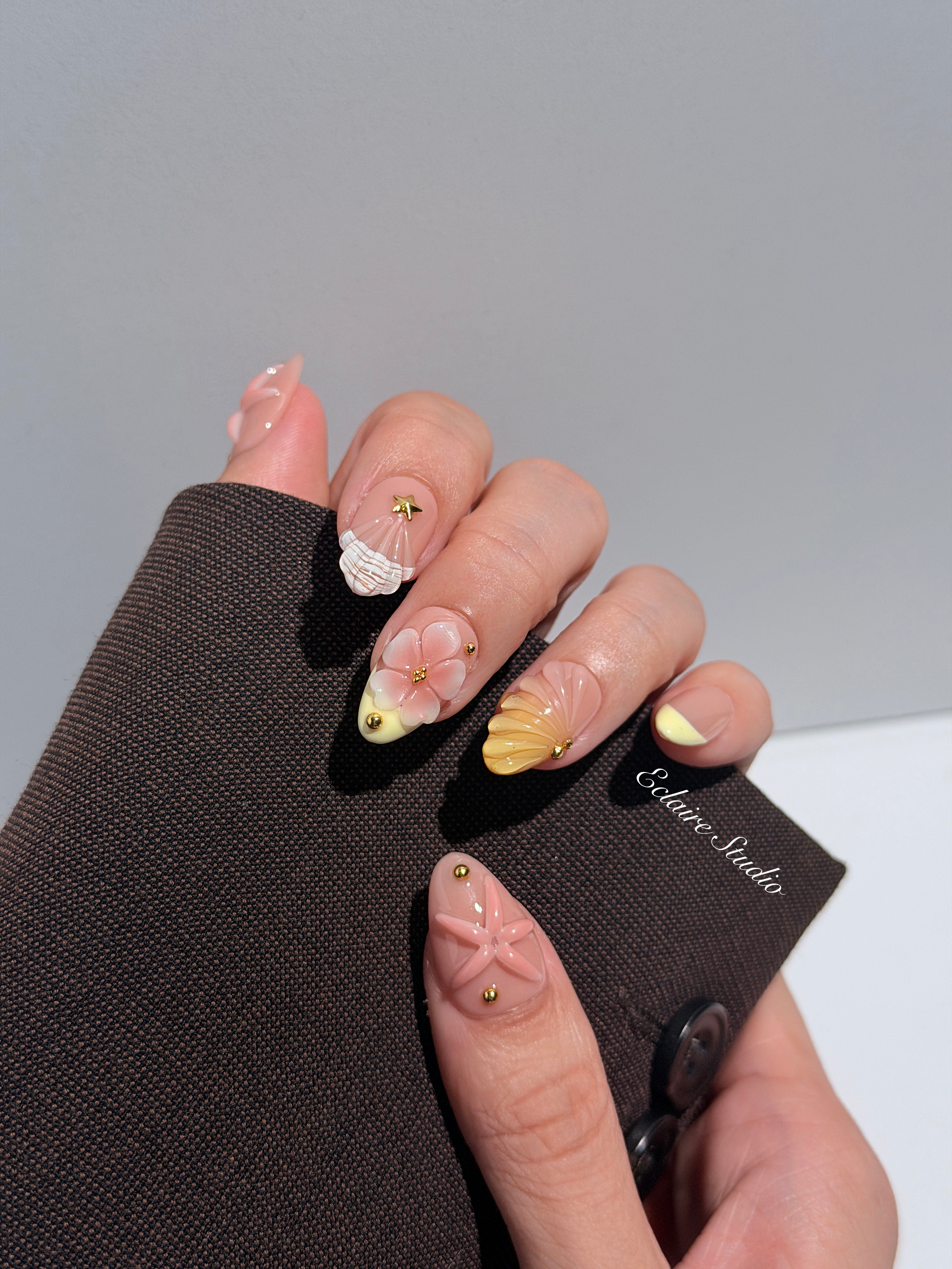 Summer Eclaire Nail Studio Luxury short almond press-on nails featuring a sheer nude base with butter yellow French tips, hand-sculpted 3D pink starfish, amber-tinted 3D seashells, and flowers with gold accents.