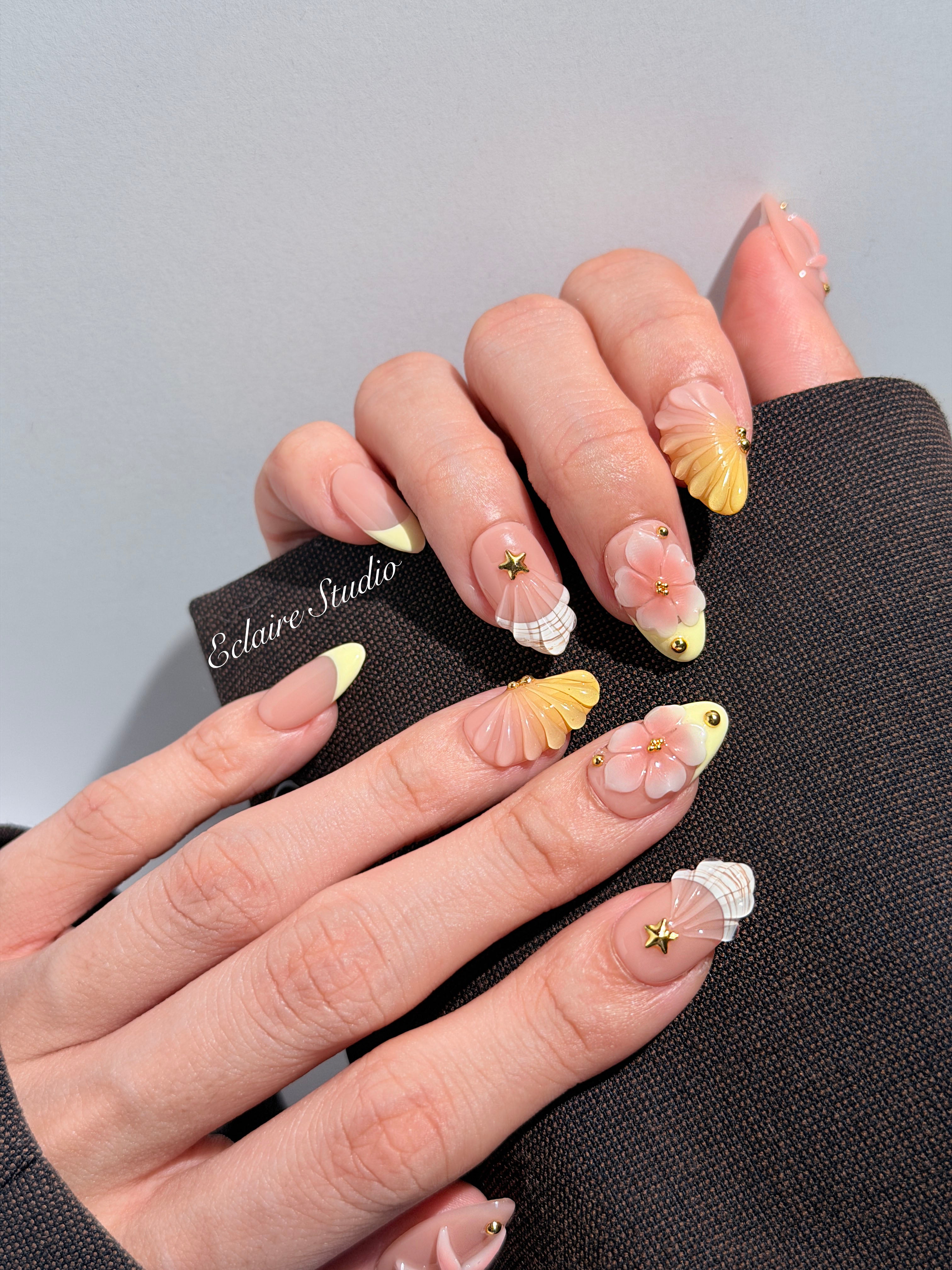 Summer Eclaire Nail Studio Luxury short almond press-on nails featuring a sheer nude base with butter yellow French tips, hand-sculpted 3D pink starfish, amber-tinted 3D seashells, and flowers with gold accents.