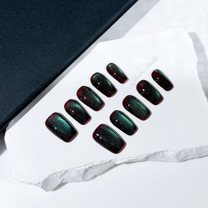 Medium coffin press-on nails with deep cherry-black base and aurora green cat-eye shimmer, arranged on a white stone surface.