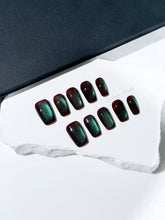 Medium coffin press-on nails with deep cherry-black base and aurora green cat-eye shimmer, arranged on a white stone surface.
