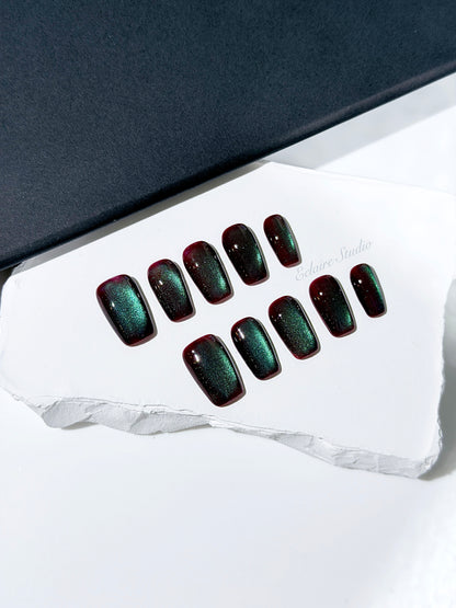 Medium coffin press-on nails with deep cherry-black base and aurora green cat-eye shimmer, arranged on a white stone surface.
