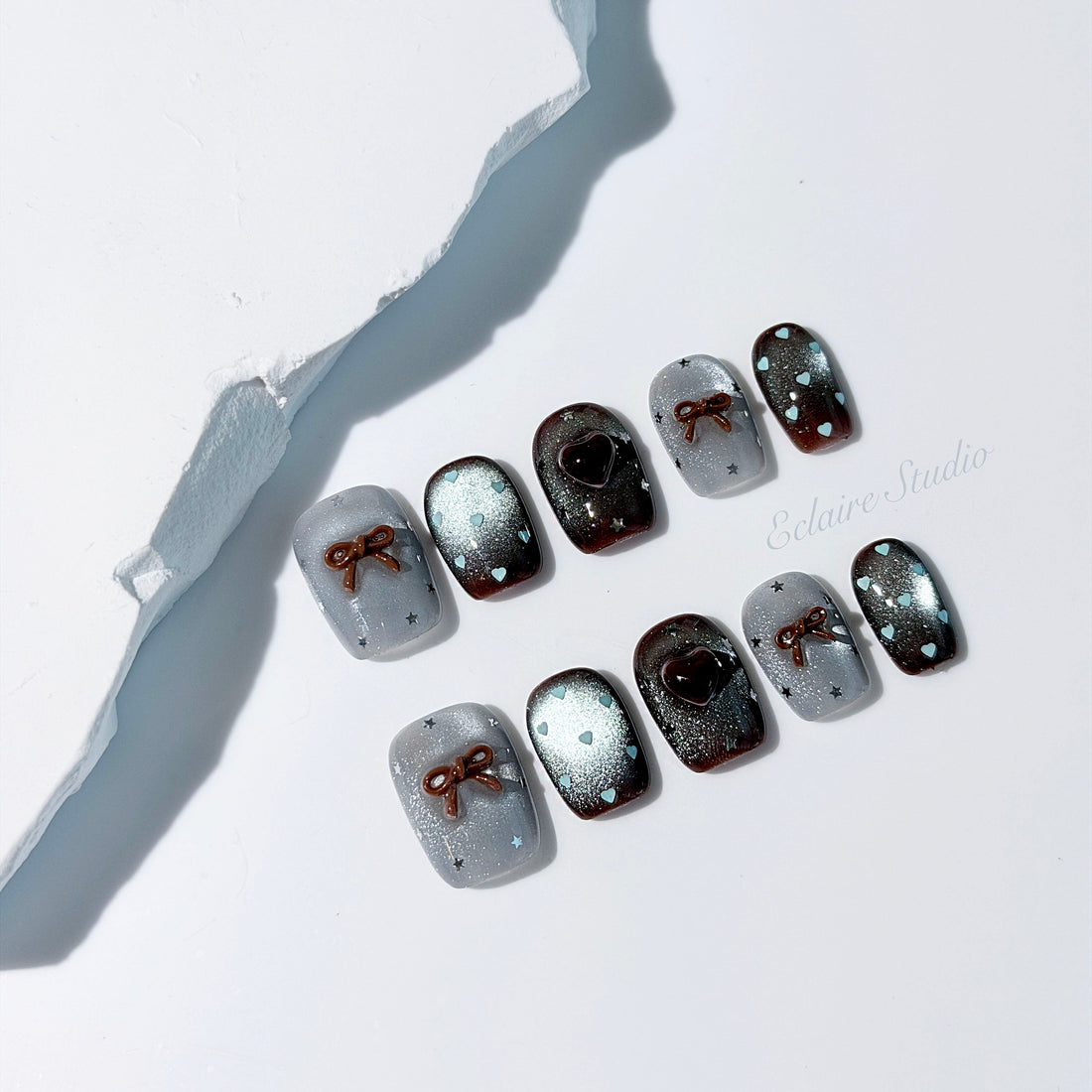 Short coffin press-on nails in mint and chocolate brown gradient with 3D heart and bow accents, displayed in a clear Éclaire Studio case under soft lighting.